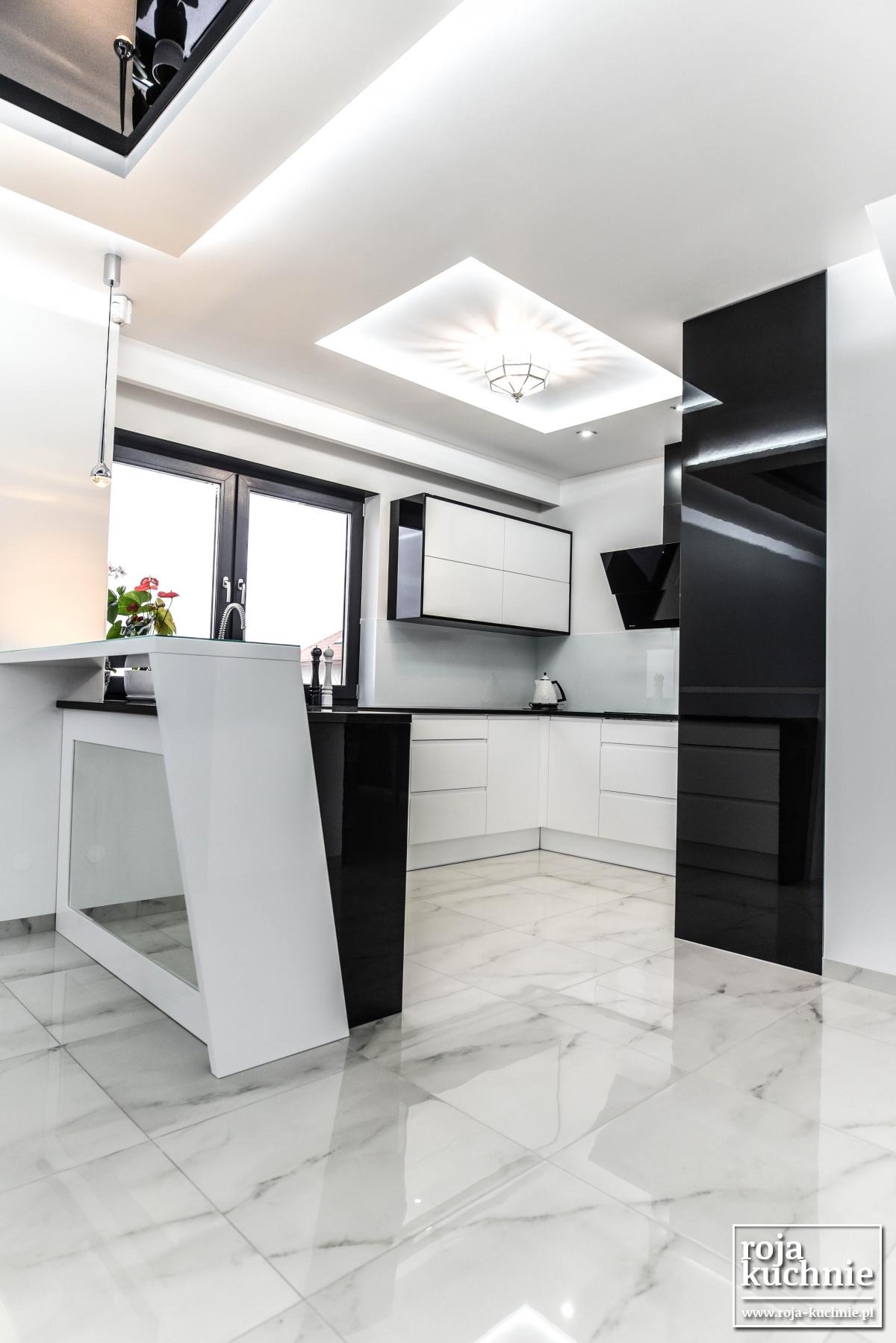 Nowoczesna, biało-czarna kuchnia z wyspą, marmurową podłogą i połyskującymi frontami szafek, oświetlona lampą wiszącą i światłem z sufitu.