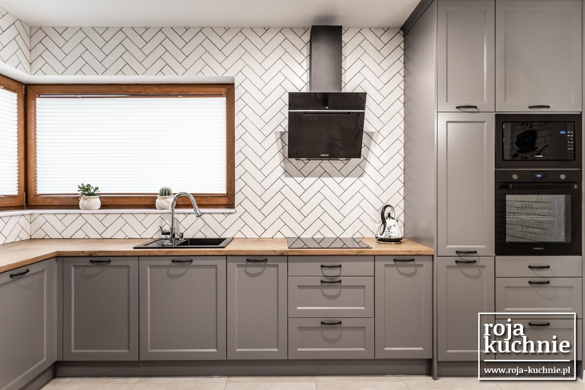 Szara zabudowa kuchenna z drewnianym blatem i białymi płytkami ułożonymi w jodełkę, czarny okap i zlew, okno z drewnianą ramą i roletami plisowanymi, czajnik w kropki.