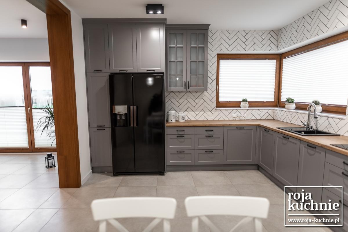 Szara zabudowa kuchenna z drewnianym blatem i czarną lodówką side-by-side, widok z perspektywy dwóch krzeseł, biała cegła na ścianie, okna z drewnianą ramą.