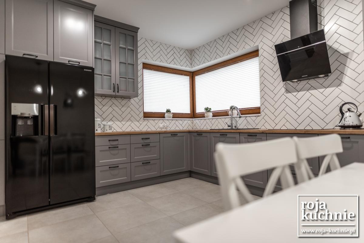 Szara kuchnia z drewnianym blatem i czarną lodówką side-by-side, białe płytki ułożone w jodełkę na ścianie, okna z drewnianą ramą i białymi żaluzjami.