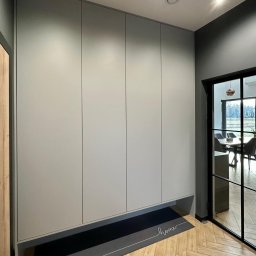 Usługi Stolarskie Ireneusz Kołodziejczyk - Szafa wnękowa w kolorze szarym z czterema drzwiami, umieszczona w nowoczesnym przedpokoju z podłogą w jodełkę i widokiem na jadalnię przez przeszklone drzwi.