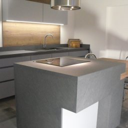 Usługi Stolarskie Ireneusz Kołodziejczyk - Nowoczesna kuchnia w Krzyżowej dolinie z szarymi frontami, drewnianym panelem nad blatem i wyspą kuchenną z płytą indukcyjną oraz stołem z krzesłami.