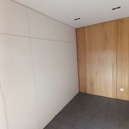 Usługi Stolarskie Ireneusz Kołodziejczyk - Wnętrze pokoju z zabudową szaf: białe szafy wnękowe po lewej, drewniane szafy po prawej, szare płytki podłogowe, oświetlenie sufitowe.