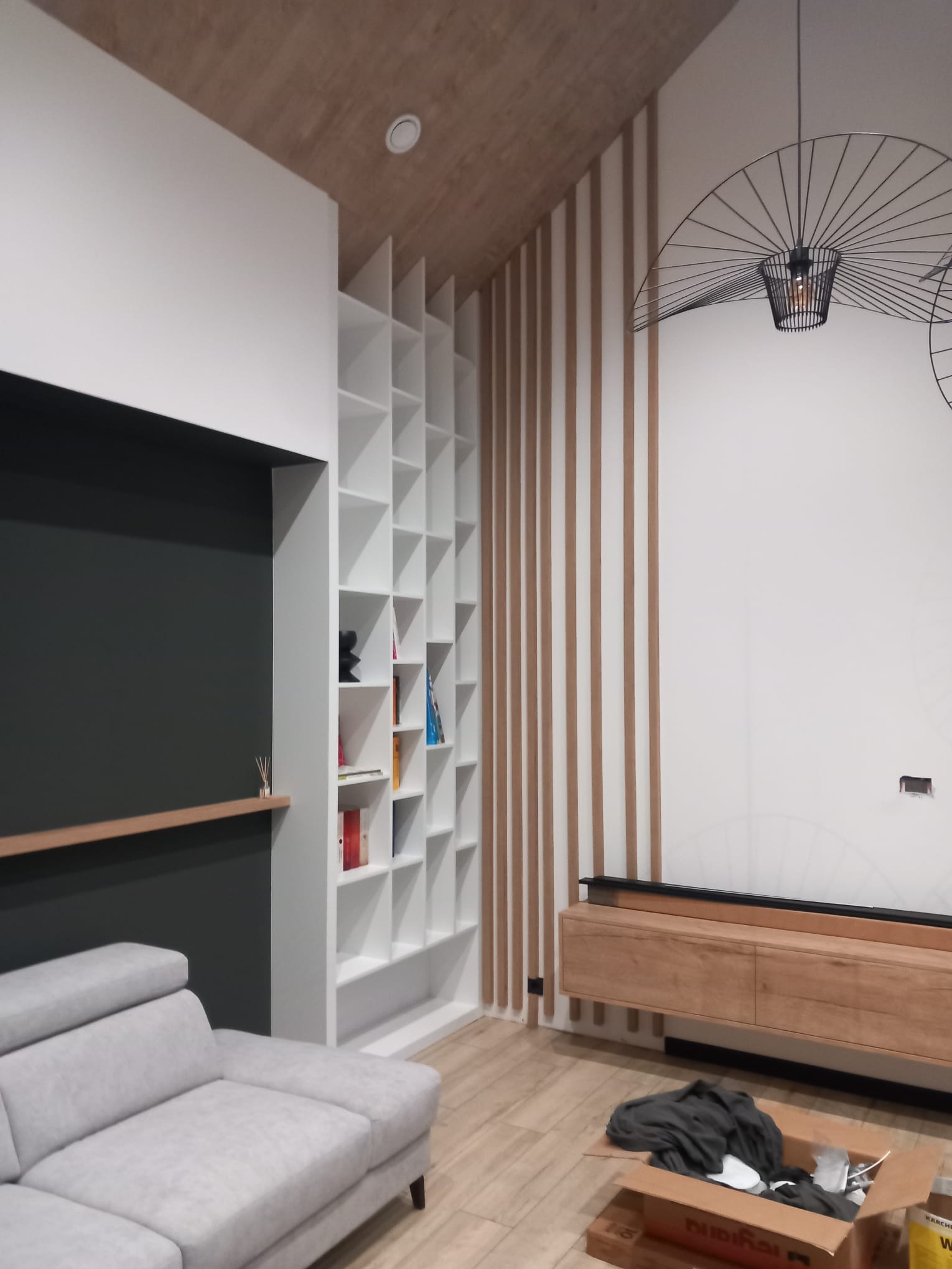 Nowoczesny salon z białą, modułową szafą wnękową na książki, drewnianymi lamelami na ścianie, szarą sofą i otwartym kartonem z akcesoriami na podłodze.