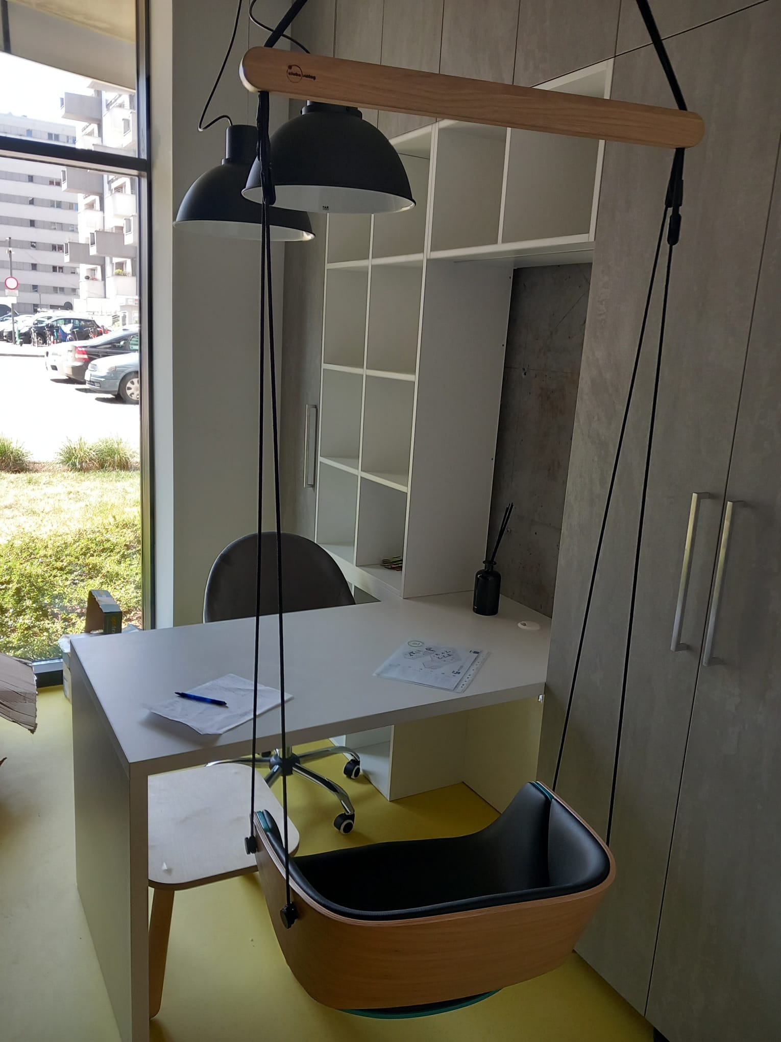 Nowoczesne biuro z wiszącym fotelem, lampą i szafą w stonowanych kolorach, widok z okna na budynki mieszkalne.