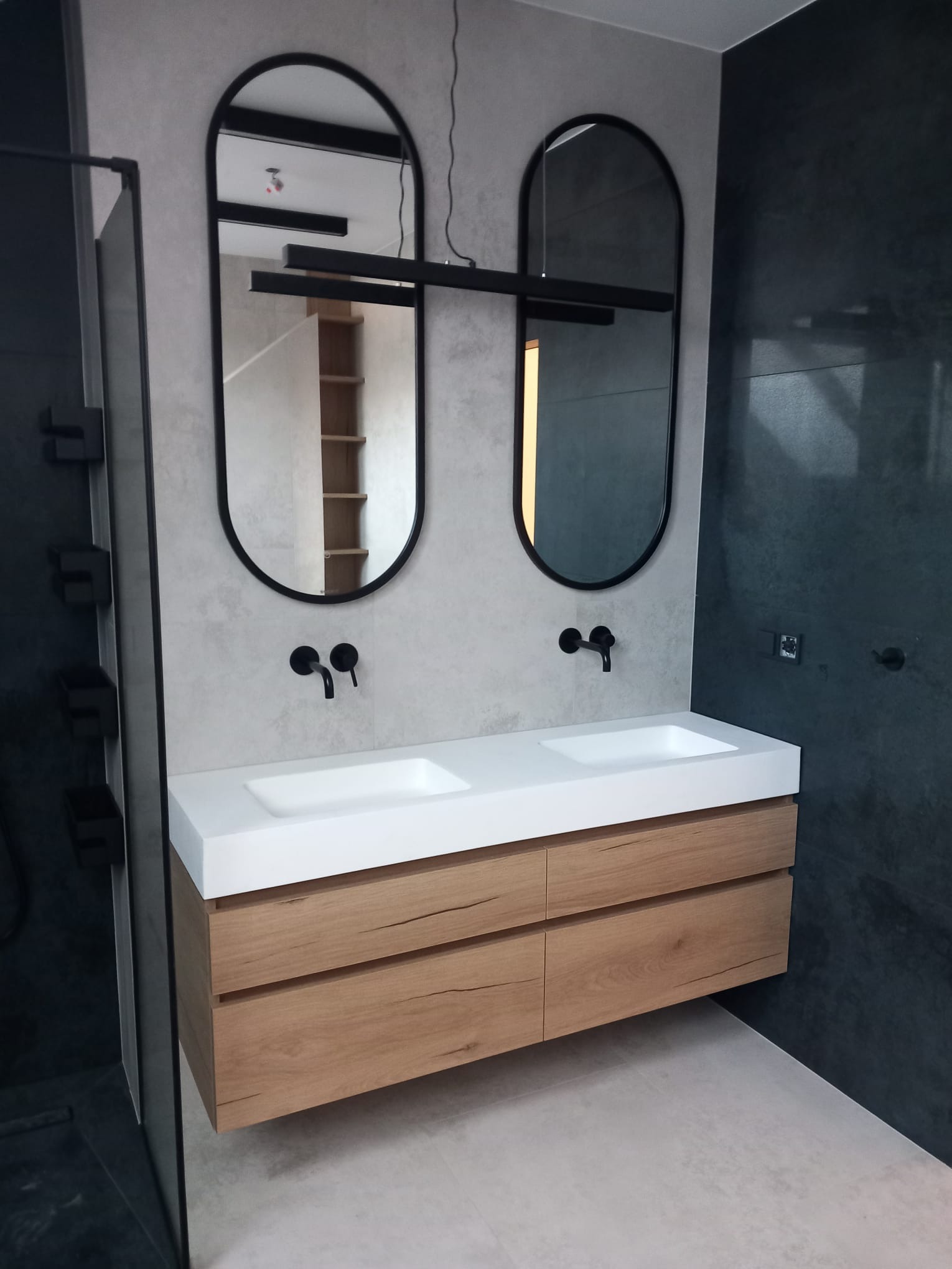Nowoczesna łazienka z podwójną umywalką, drewniana szafka wisząca, czarne baterie i owalne lustra z czarnymi ramami, oświetlenie ledowe, ściana z ciemnoszarego kamienia.