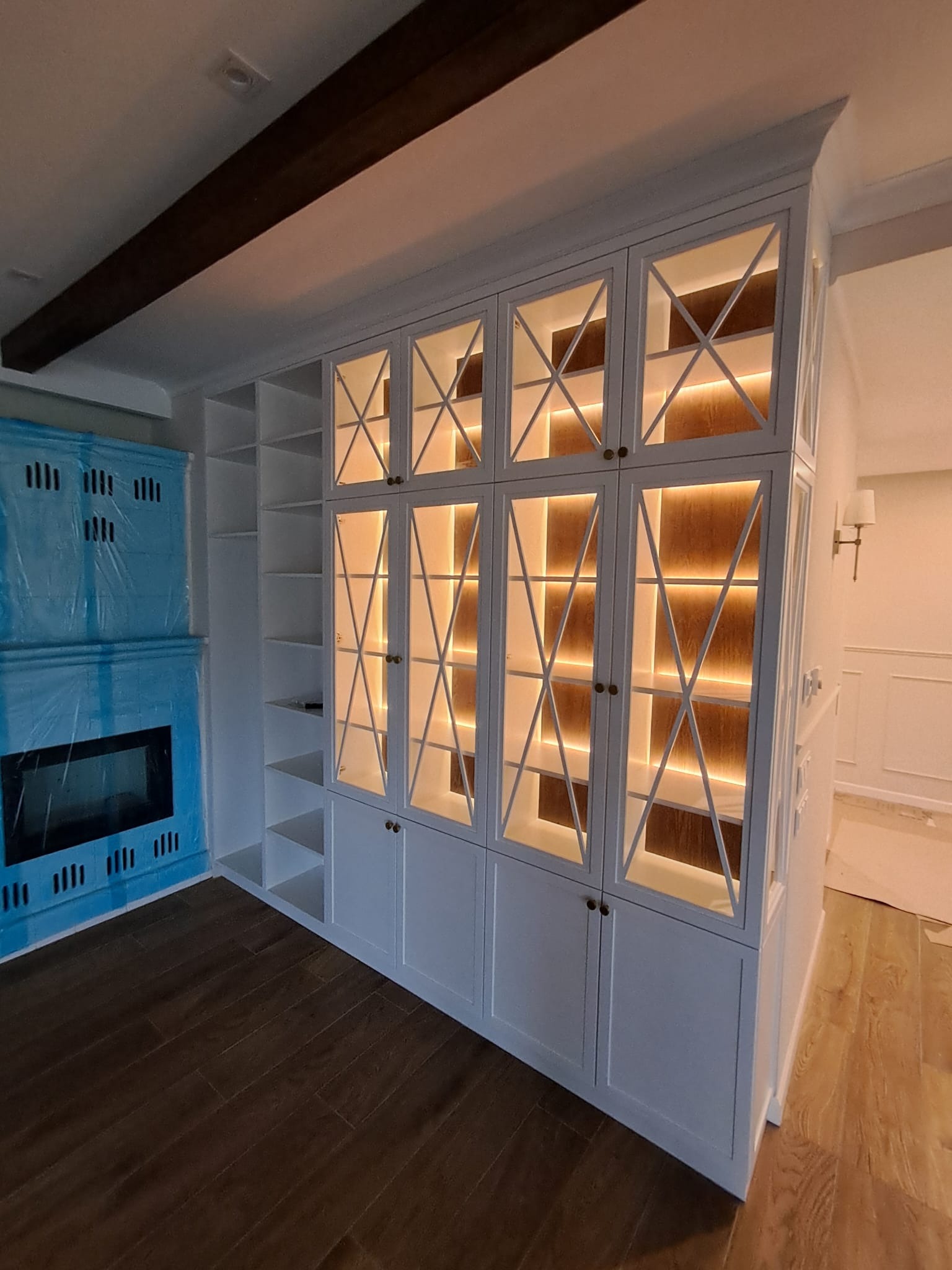 Biała szafa wnękowa z przeszklonymi drzwiami, podświetlona od wewnątrz, obok niebieskiego pieca kaflowego w nowoczesnym wnętrzu.