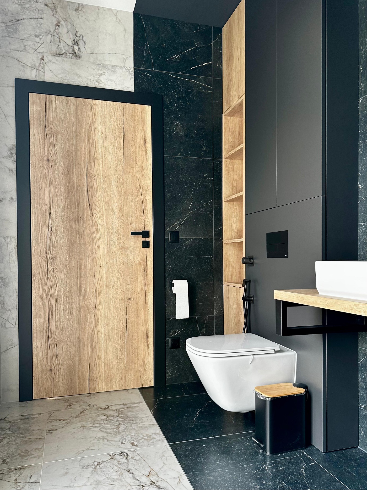 Nowoczesna łazienka z drewnianą okleiną drzwi i szafek, czarne płytki marmurowe i biała ceramika sanitarna, minimalistyczny design.