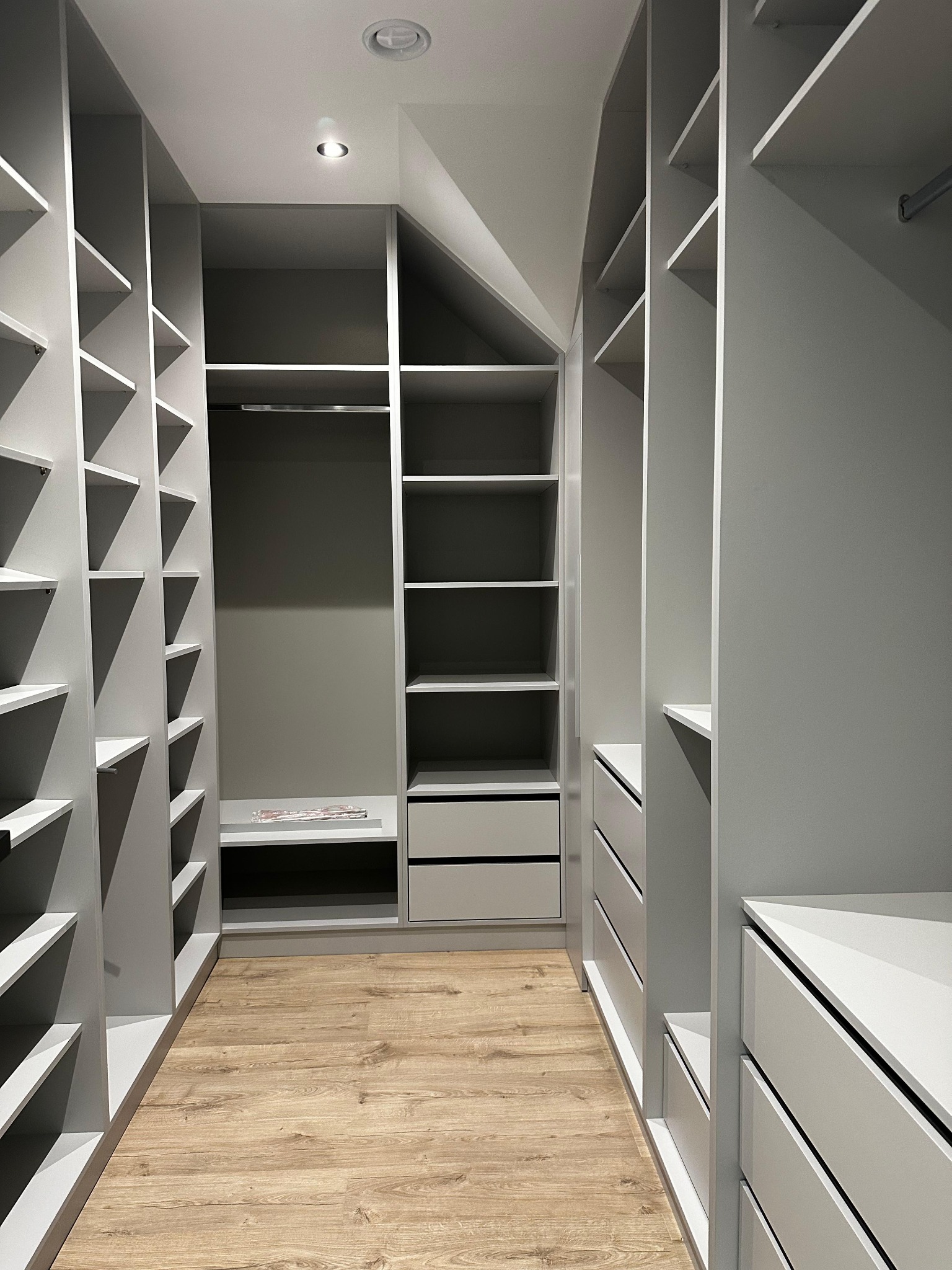 Wnętrze garderoby z szarymi szafami wnękowymi z półkami i szufladami, oświetlenie sufitowe, panele podłogowe imitujące drewno.
