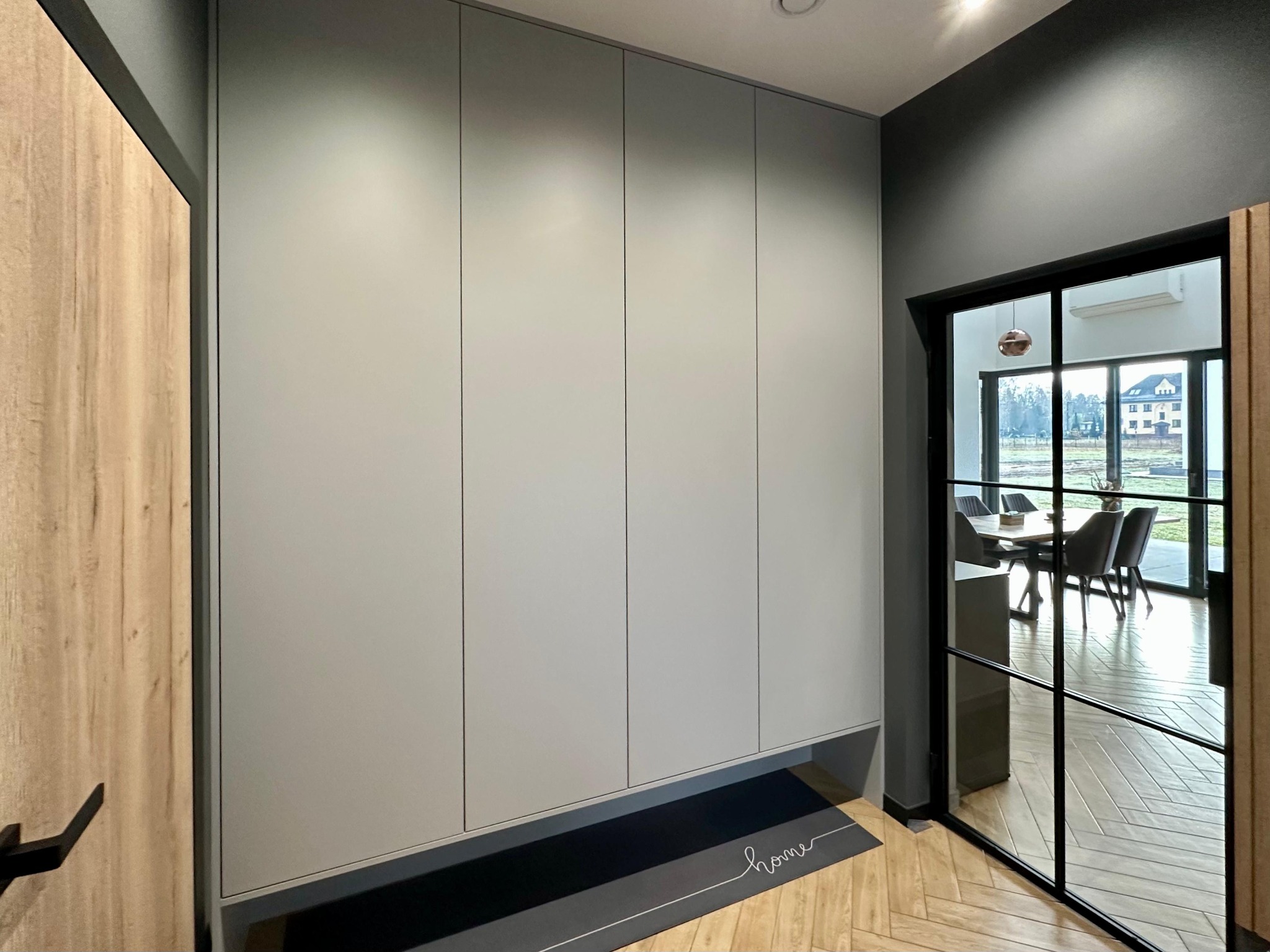 Szafa wnękowa w kolorze szarym z czterema drzwiami, umieszczona w nowoczesnym przedpokoju z podłogą w jodełkę i widokiem na jadalnię przez przeszklone drzwi.