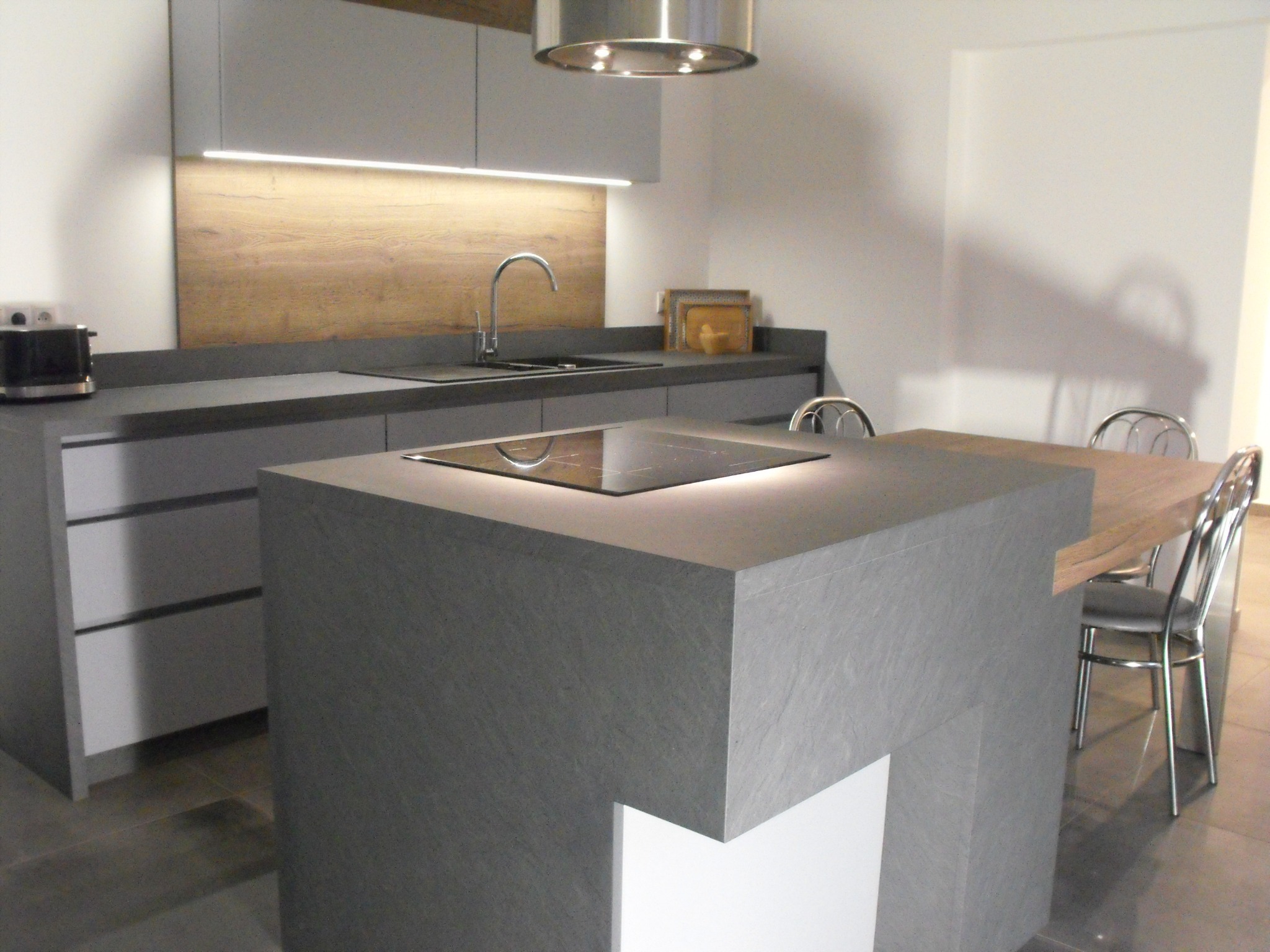 Nowoczesna kuchnia w Krzyżowej dolinie z szarymi frontami, drewnianym panelem nad blatem i wyspą kuchenną z płytą indukcyjną oraz stołem z krzesłami.