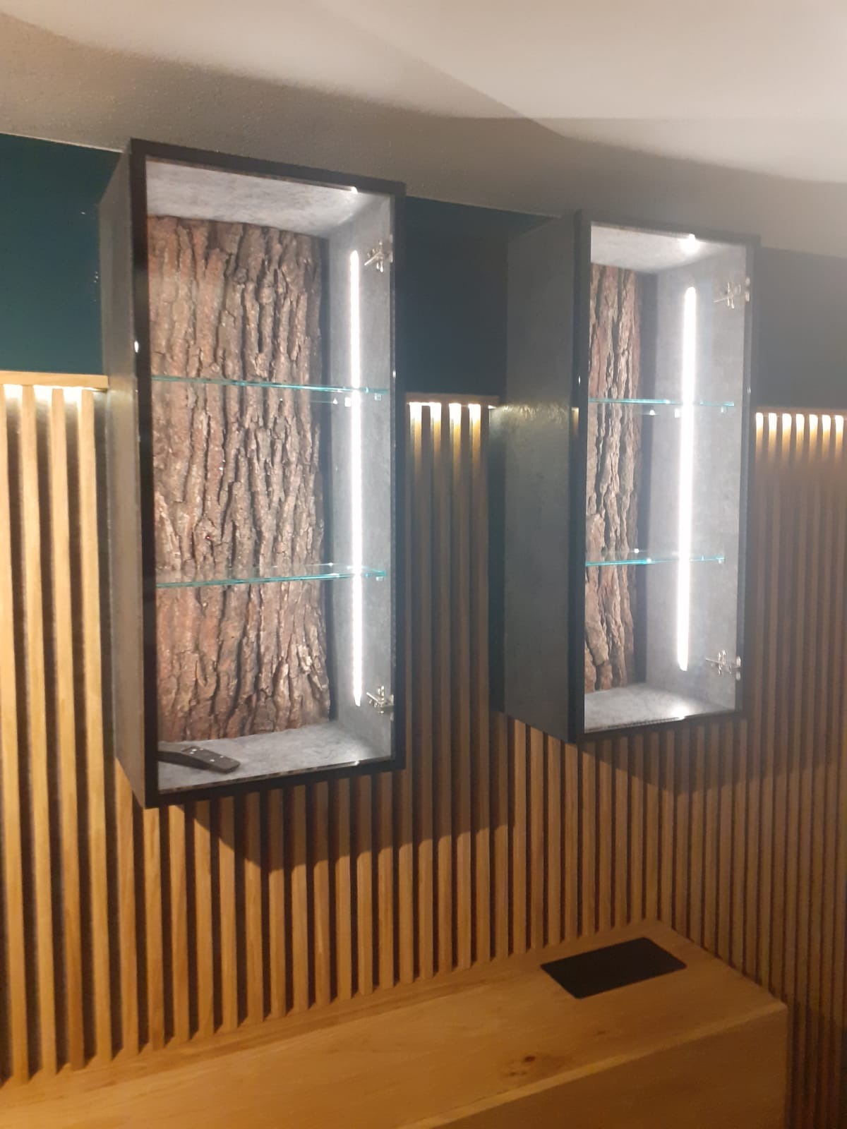 Dwie podświetlane szafki wiszące z szarymi ramami, wewnątrz imitacja pnia drzewa i szklane półki, zamontowane na ścianie z drewnianych listewek.