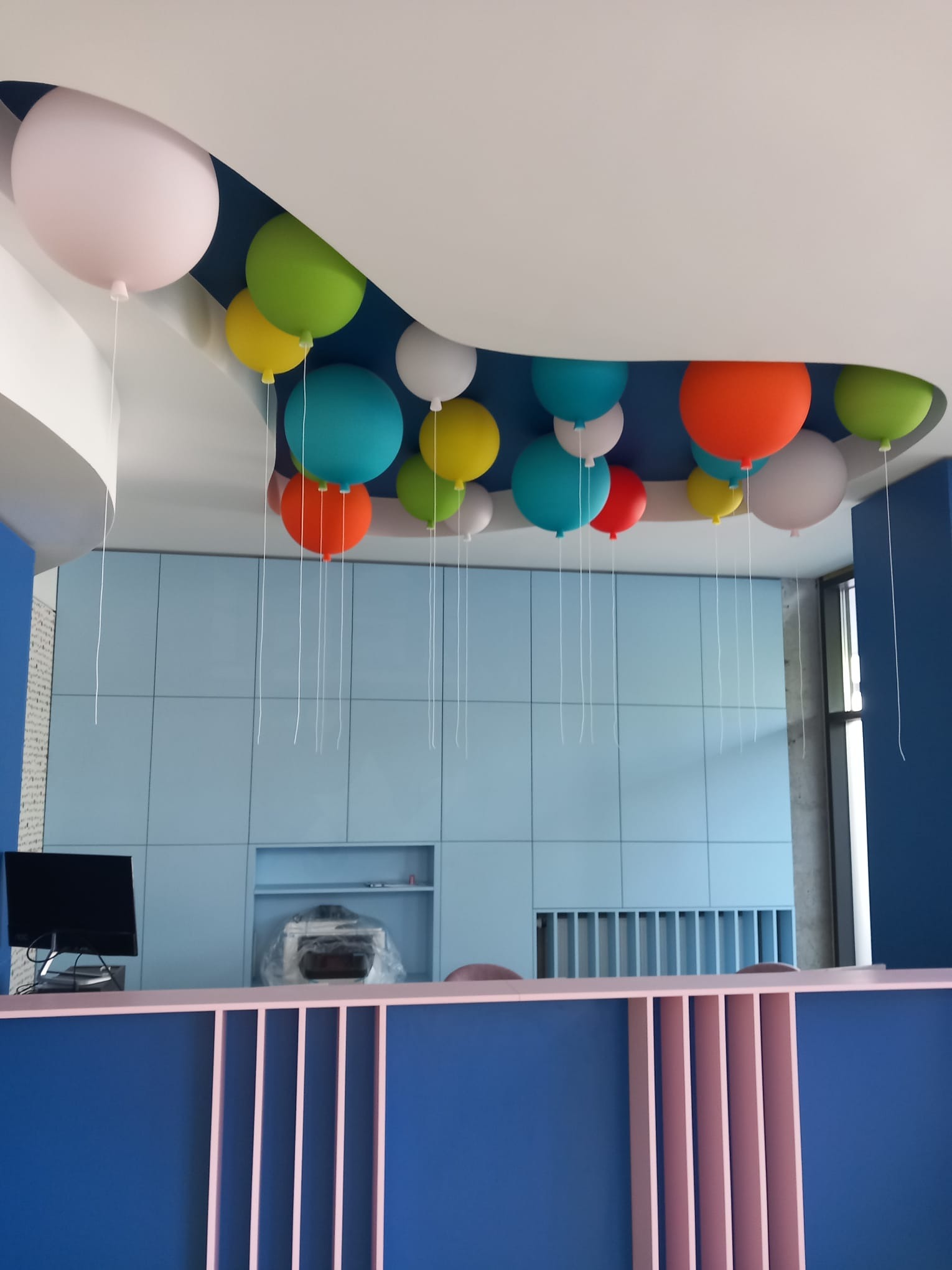 Niebieska zabudowa meblowa z pastelowymi balonami pod sufitem, widoczny blat z różowymi pionowymi listwami, monitor i zapakowane urządzenie na blacie.