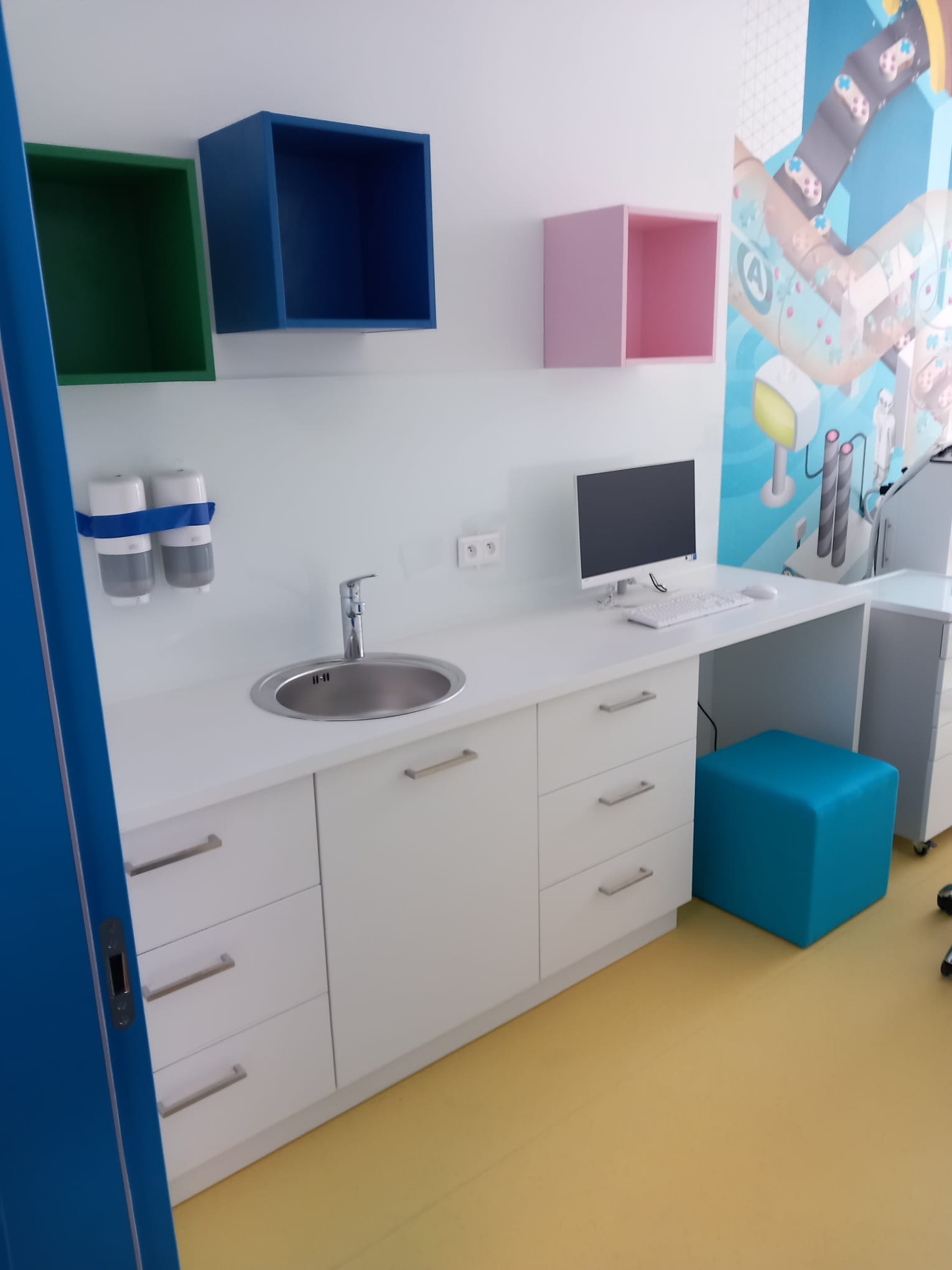 Biała zabudowa meblowa z umywalką i blatem biurkowym, kolorowe szafki wiszące, grafika na ścianie, niebieski puf i żółta podłoga w pokoju dziecięcym.