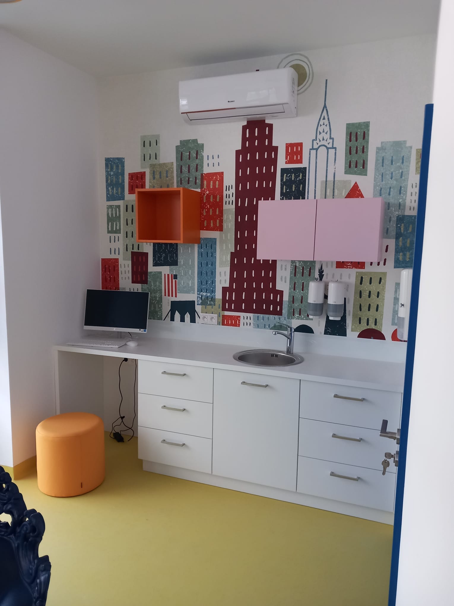 Białe szafki z szufladami pod blatem z umywalką, pomarańczowa otwarta szafka wisząca i różowa szafka wisząca na ścianie z motywem miasta, klimatyzator na górze, żółta podłoga i pomarańczowy puf.
