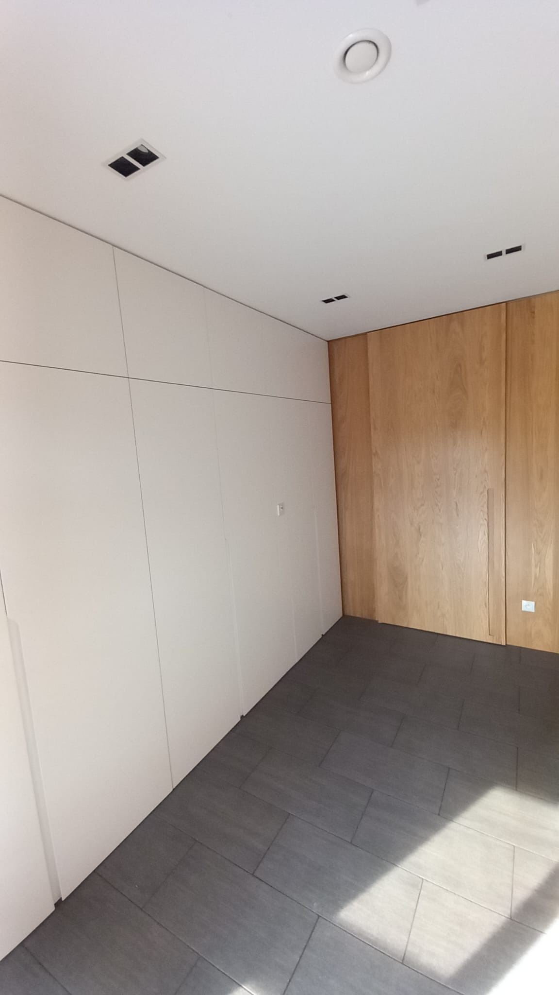 Wnętrze pokoju z zabudową szaf: białe szafy wnękowe po lewej, drewniane szafy po prawej, szare płytki podłogowe, oświetlenie sufitowe.