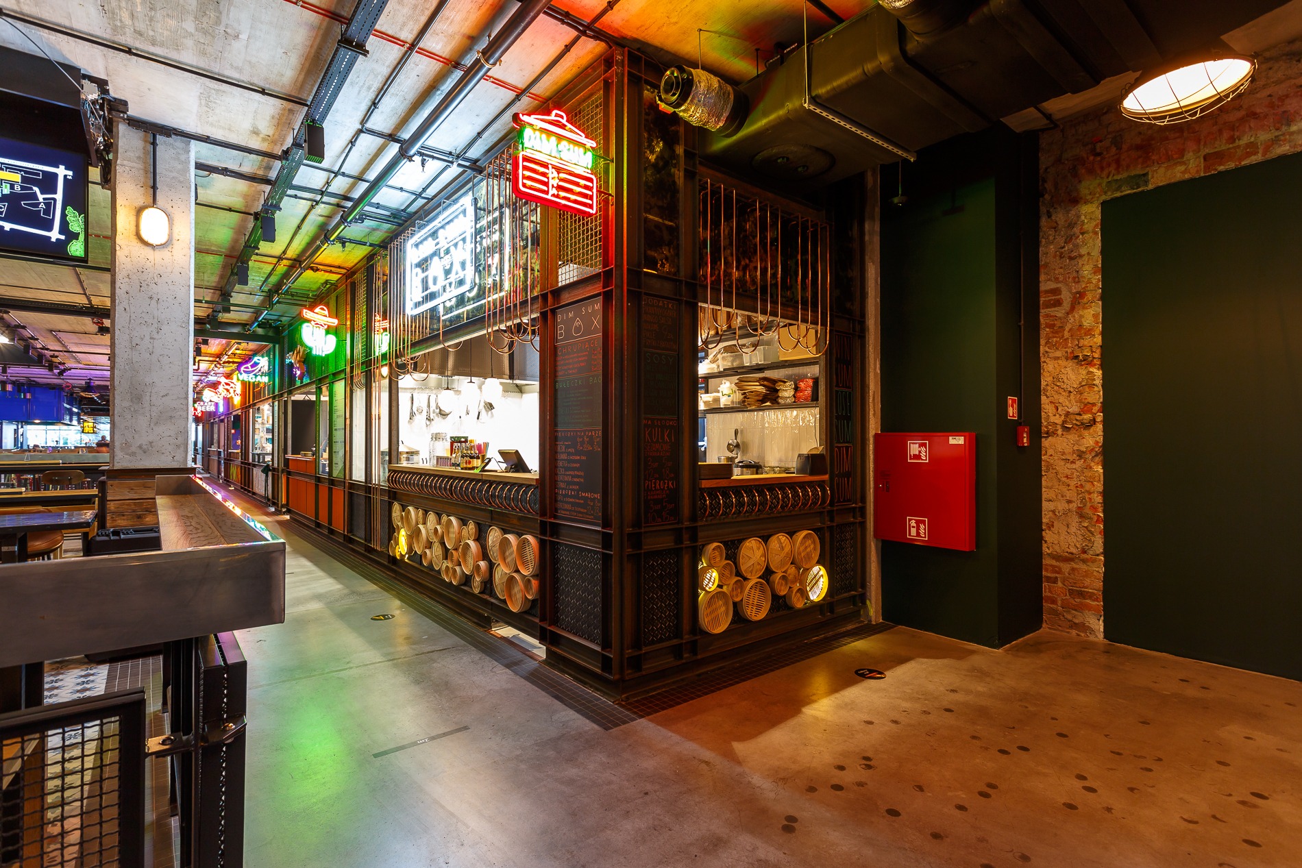 Wnętrze lokalu gastronomicznego z otwartą kuchnią i neonowymi szyldami, widok z perspektywy klienta, surowy betonowy sufit z widocznymi instalacjami, dekoracyjne okręgi z podświetleniem na froncie...