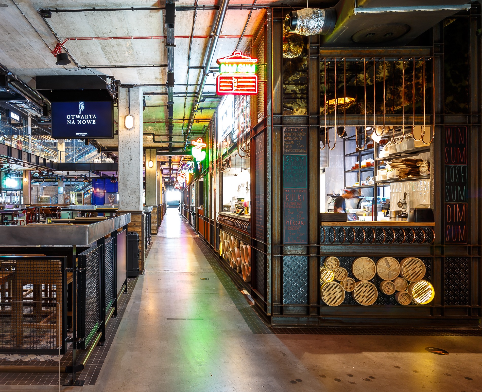 Wnętrze hali gastronomicznej z rzędem stoisk z jedzeniem, neonowe szyldy nad stoiskami, dekoracyjne elementy z bambusowych koszyczków, betonowa podłoga z refleksami kolorowego światła.