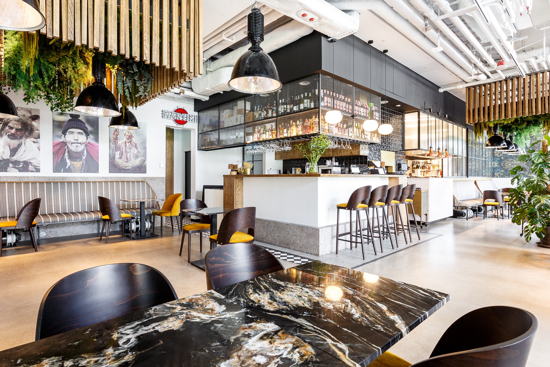 Wnętrze restauracji w stylu industrialnym z marmurowymi stolikami, drewnianymi krzesłami z żółtymi siedziskami, barem z metalową siatką i wiszącymi lampami oraz dekoracjami roślinnymi.