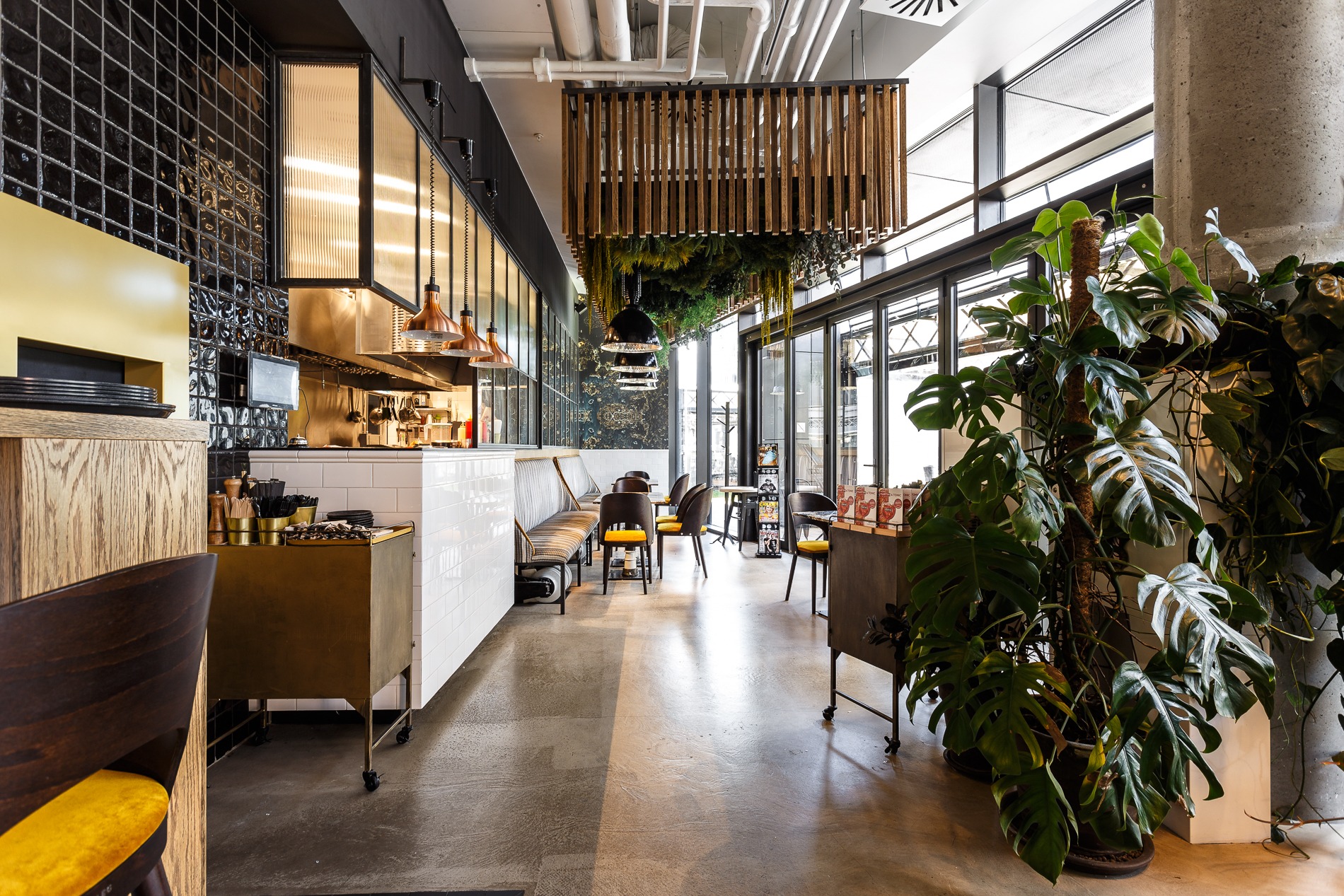 Nowoczesne wnętrze restauracji z otwartą kuchnią, drewnianą konstrukcją sufitową z zielenią, betonowym filarem i dużymi oknami, z widocznymi elementami wykończenia ścian i podłóg.