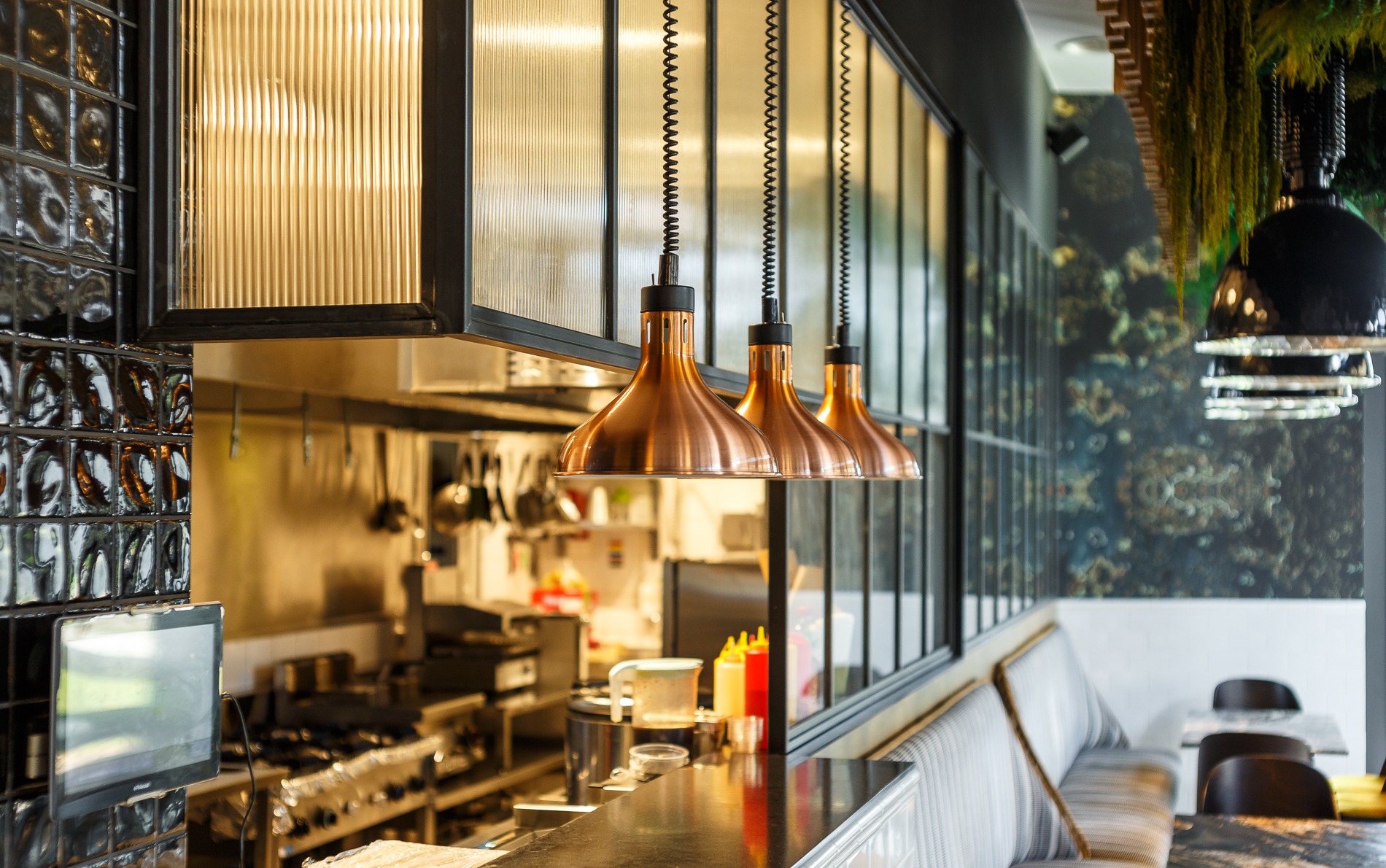 Wnętrze restauracji z miedzianymi lampami wiszącymi nad barem, czarne płytki na ścianie i widok na kuchnię w tle.