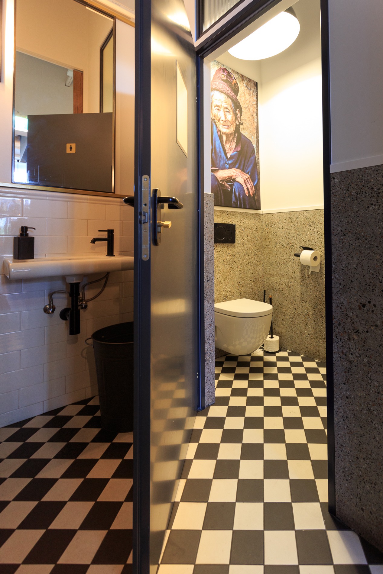 Nowoczesna łazienka z czarno-białą szachownicą na podłodze, widoczna umywalka z czarną armaturą, lustro z podświetleniem oraz toaleta z portretem starszej kobiety na ścianie.