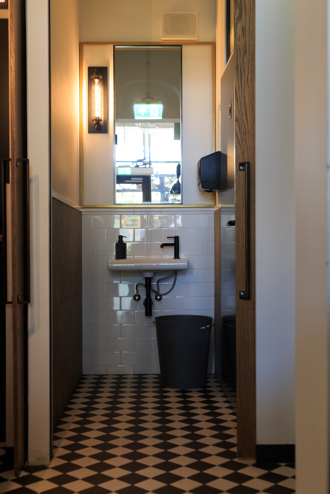 Wnętrze toalety w Warszawie z białą umywalką, czarną armaturą i koszem na śmieci na podłodze w szachownicę, widok z korytarza.