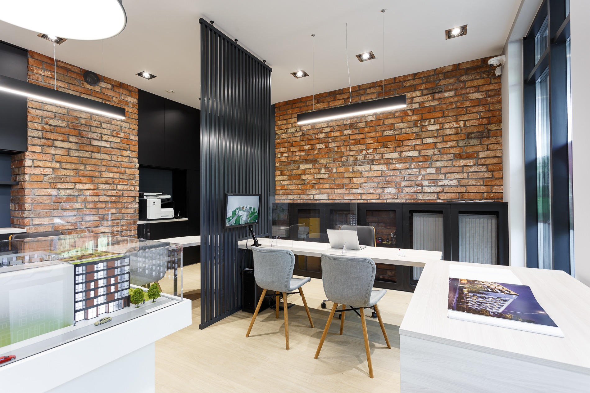 Wnętrze biura nieruchomości z biurkiem, krzesłami, ścianą z cegły i modelem architektonicznym budynku.