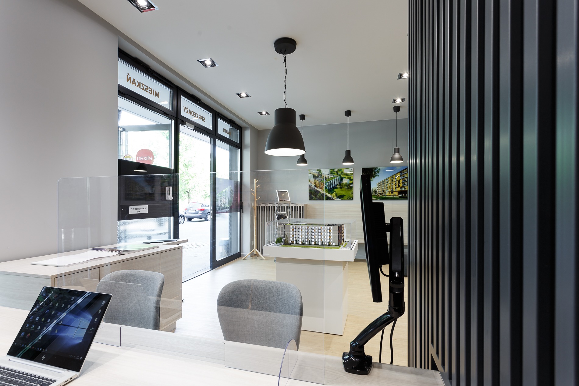 Wnętrze biura sprzedaży mieszkań z modelem architektonicznym budynku na białym podeście, monitor na regulowanym ramieniu, szare krzesła i przezroczyste osłony biurkowe, widok na ulicę przez...