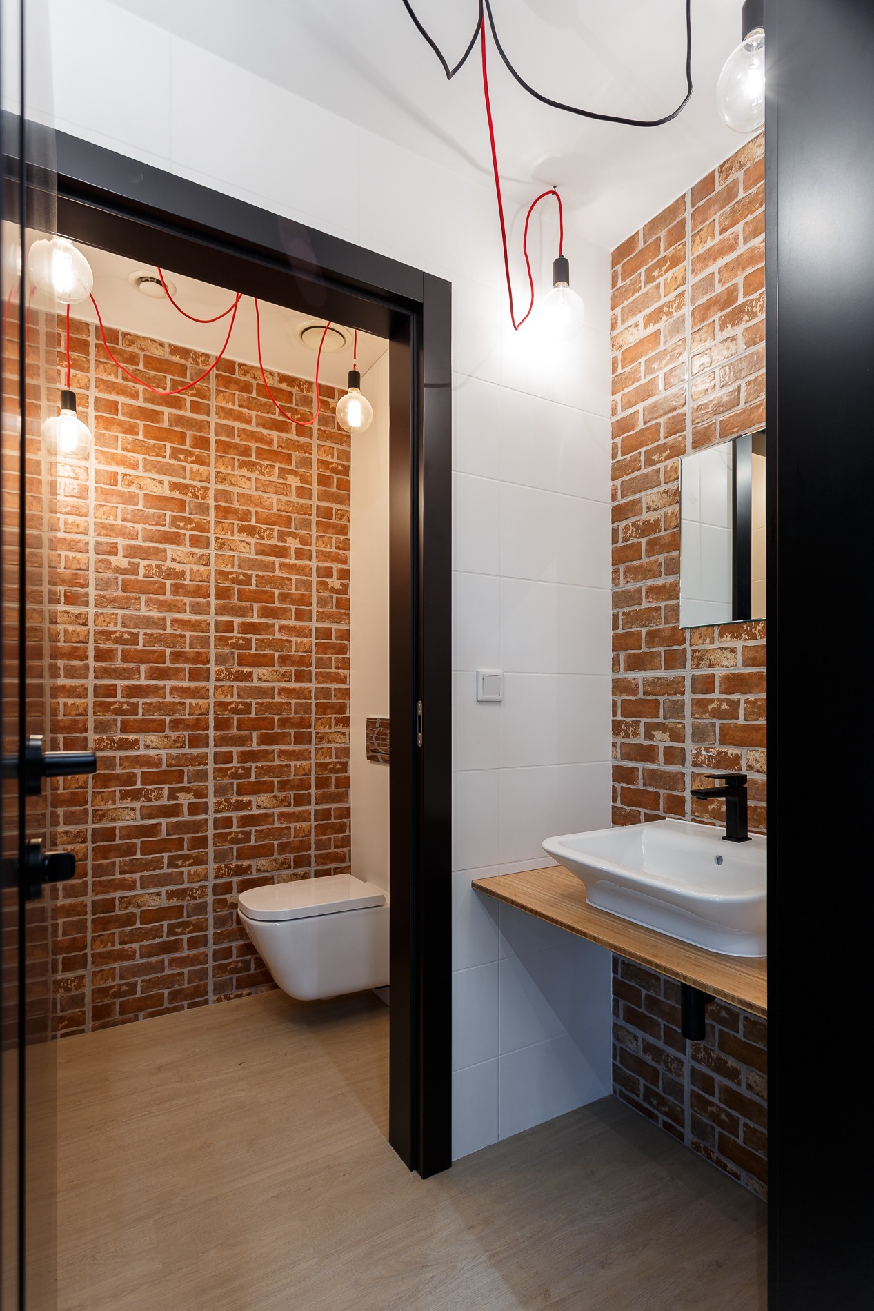 Nowoczesna łazienka z ceglaną ścianą, wiszącą toaletą i umywalką na drewnianym blacie, oświetlona wiszącymi żarówkami z czerwonymi kablami.