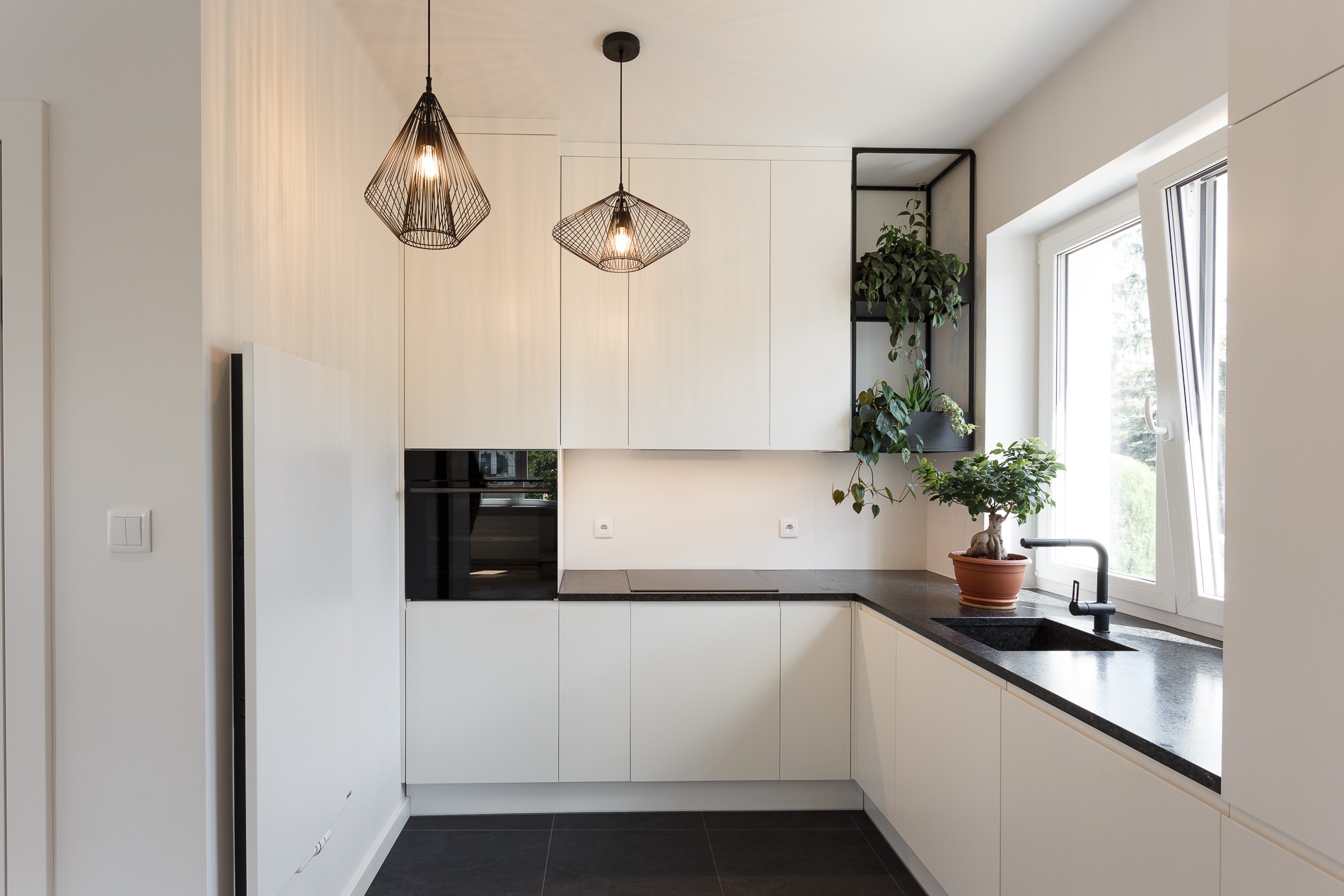 Nowoczesna kuchnia z białymi szafkami, czarnym blatem i podłogą, oświetlona wiszącymi lampami w kształcie klatek i doświetlona światłem dziennym z okna z widokiem na zieleń. Na blacie stoi bonsai...