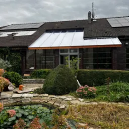 Zadaszenie tarasu z poliwęglanu przy domu z cegły, widok na ogród z oczkiem wodnym i kwiatami, pochmurne niebo.