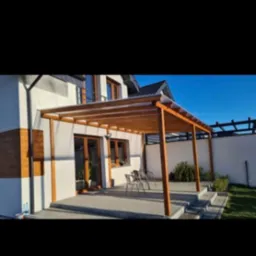 Nowoczesna drewniana konstrukcja z przezroczystym zadaszeniem, przylegająca do białego domu z brązowymi ramami okiennymi, widoczne krzesła ogrodowe i roślina w donicy.