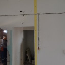 Kacper Stańczyk - Wnętrze w trakcie remontu z widocznymi przewodami elektrycznymi poprowadzonymi po ścianach i suficie, zabezpieczonymi żółtą taśmą, oraz fragmentem osoby w pomarańczowych rękawicach w przejściu.