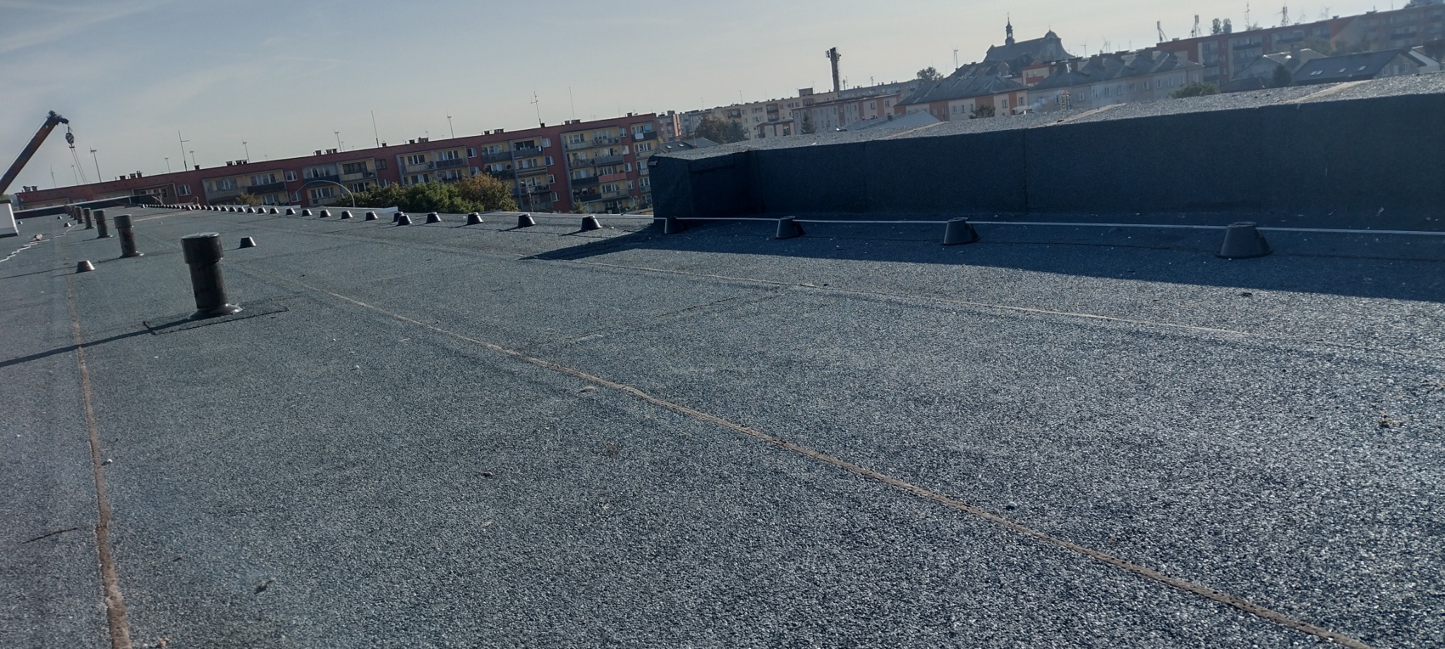 Widok z góry na płaski dach pokryty szarym, bitumicznym materiałem z widocznymi odpowietrznikami i wspornikami pod instalacje, na tle budynków miejskich i dźwigu budowlanego.