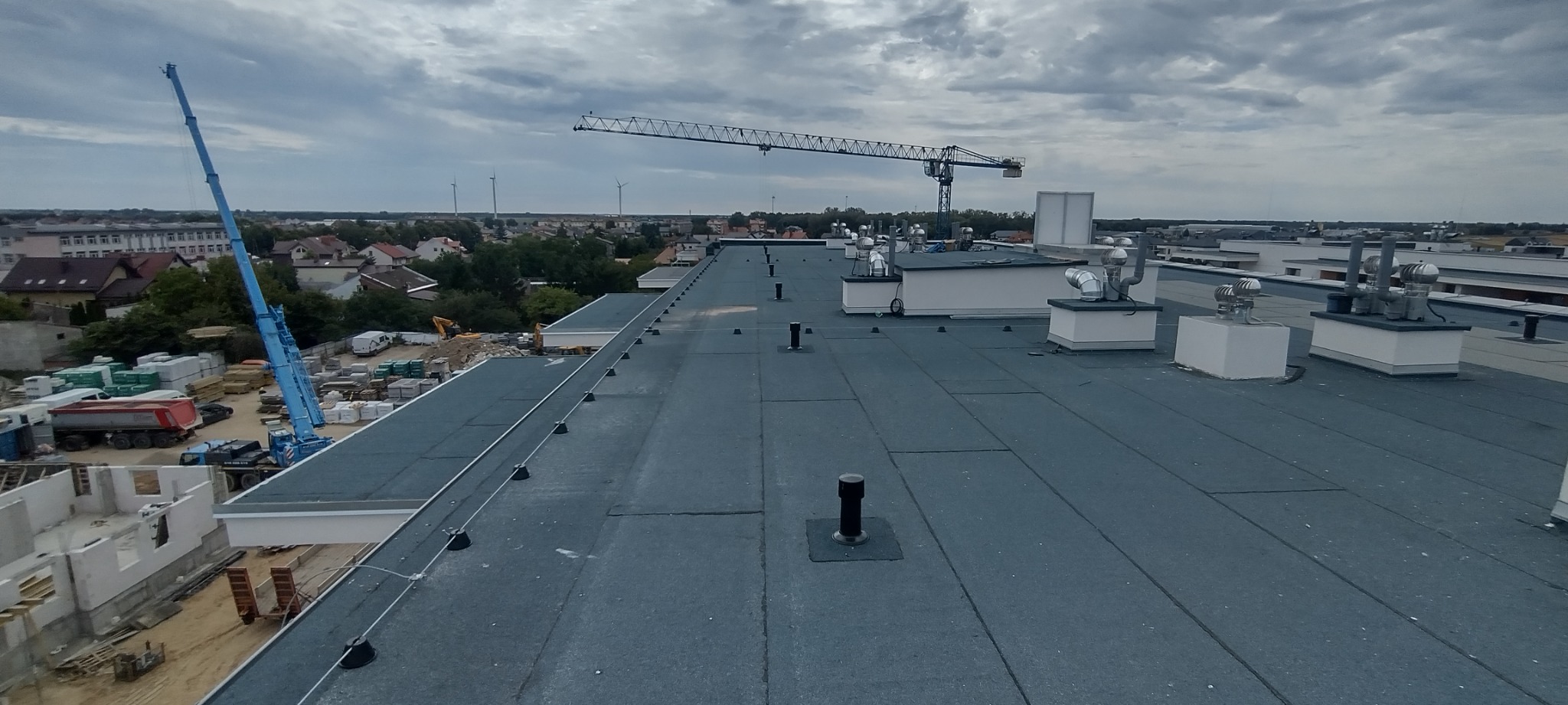 Rozległy, płaski dach pokryty papą termozgrzewalną z widocznymi kominkami wentylacyjnymi i odgromnikami, w tle budowa z dźwigami i zabudowa miejska pod pochmurnym niebem.