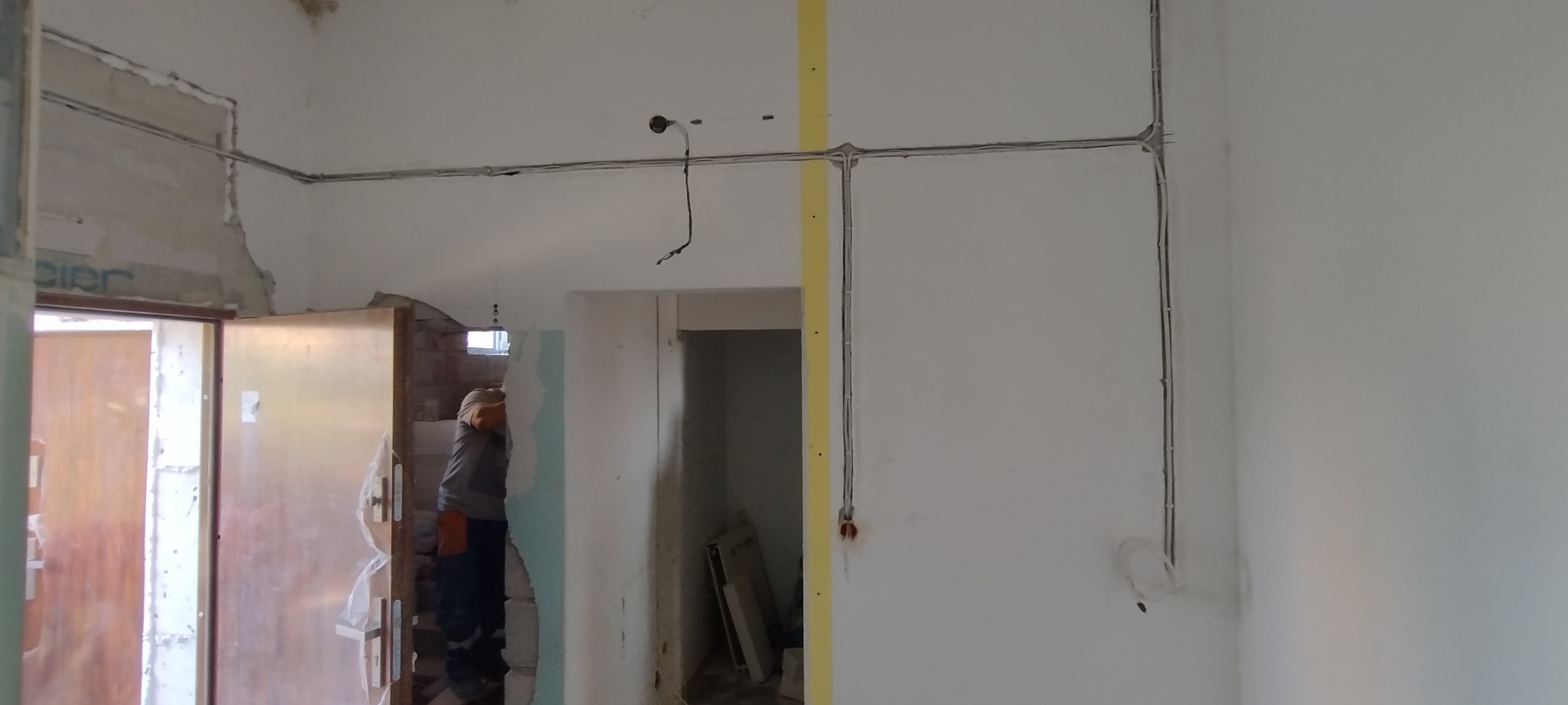 Wnętrze w trakcie remontu z widocznymi przewodami elektrycznymi poprowadzonymi po ścianach i suficie, zabezpieczonymi żółtą taśmą, oraz fragmentem osoby w pomarańczowych rękawicach w przejściu.