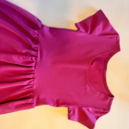 Dziewczęca sukienka w kolorze fuksji z krótkim rękawem i marszczeniem w talii, leżąca na beżowym tle, widok z góry.