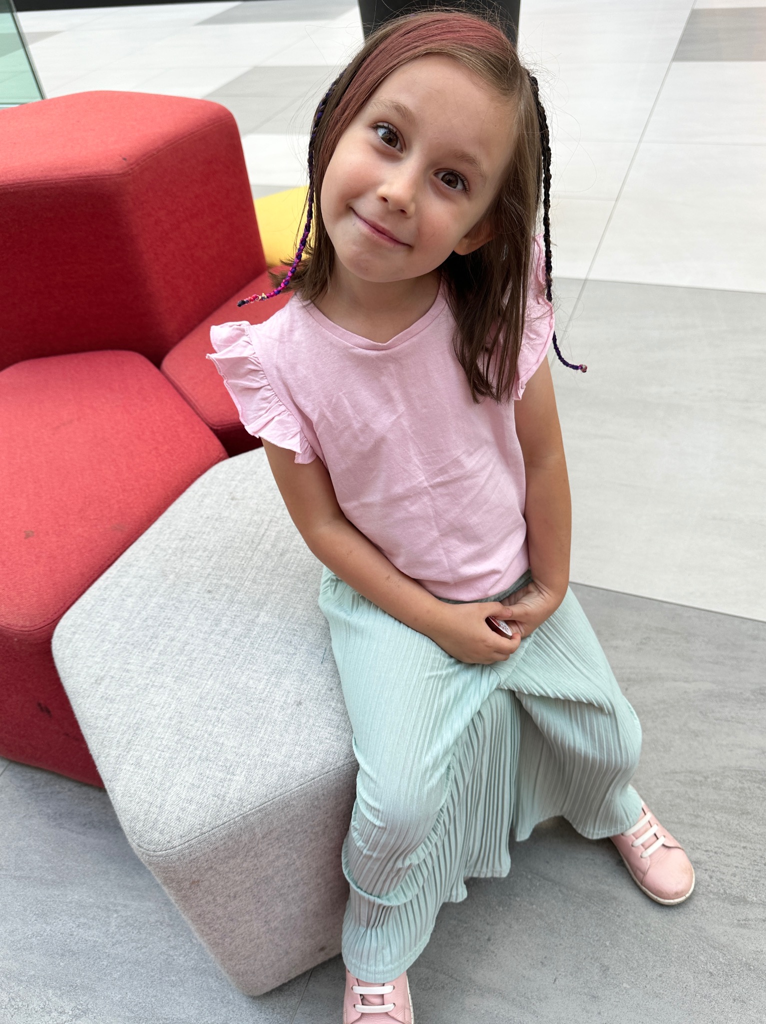 Dziewczynka w różowej bluzce i plisowanych, miętowych spodniach siedzi na szarym pufie, obok czerwonych pufów, uśmiechając się do kamery.
