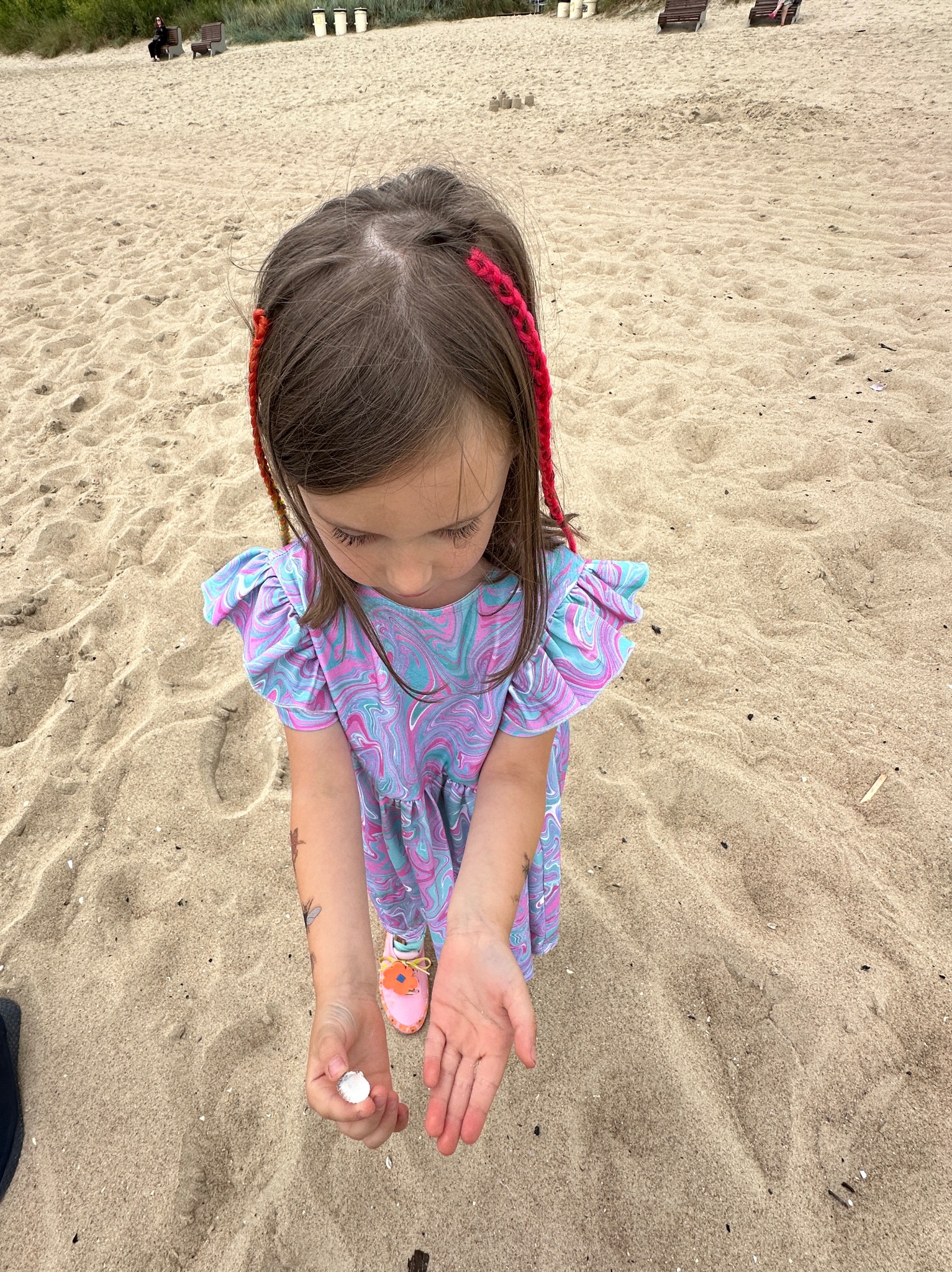 Dziewczynka w sukience z marmurkowym wzorem prezentuje znalezioną muszelkę na piaszczystej plaży, z widocznymi detalami warkoczy i tatuaży tymczasowych na ramionach.
