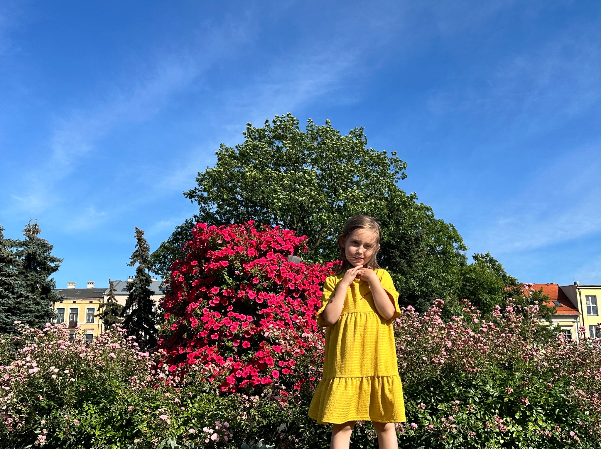 Dziewczynka w żółtej sukience w ogrodzie z kwitnącymi różowymi i czerwonymi kwiatami w słoneczny dzień.