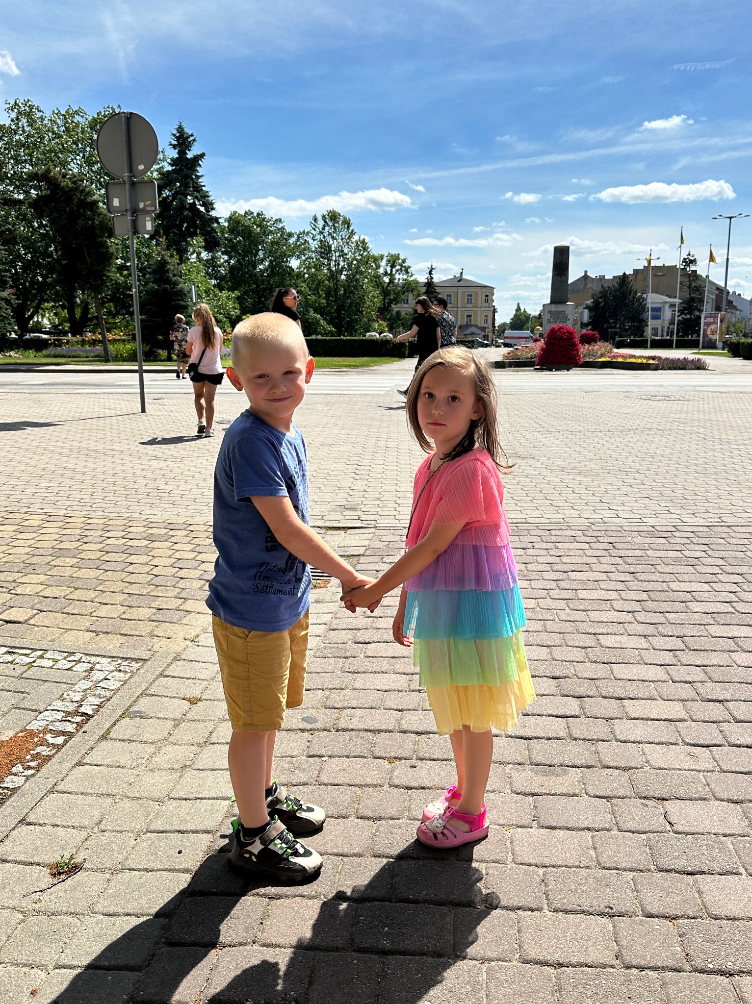 Uśmiechnięte dzieci trzymają się za ręce na tle miejskiego placu; dziewczynka w tęczowej sukience z falbankami.