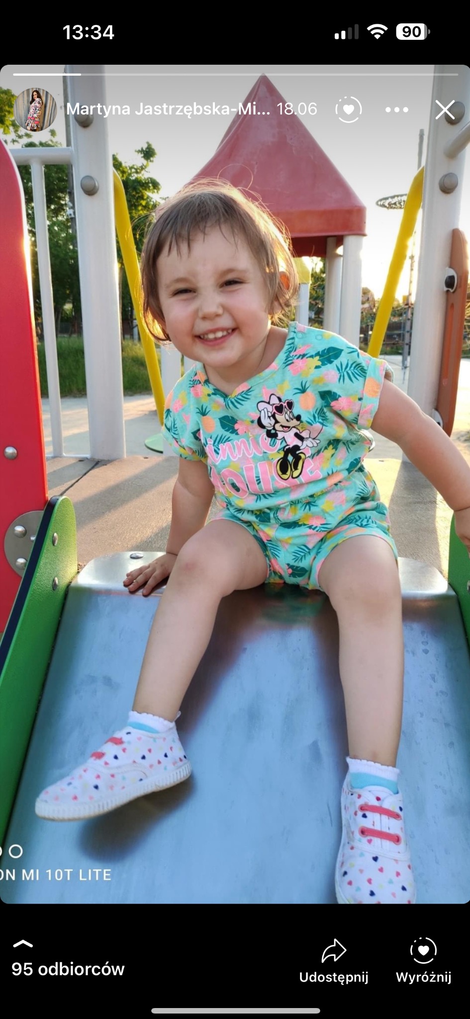 Uśmiechnięta dziewczynka w komplecie z krótkim rękawem i szortami w tropikalny wzór z Minie Mouse siedzi na metalowej zjeżdżalni na placu zabaw, prezentując strój.