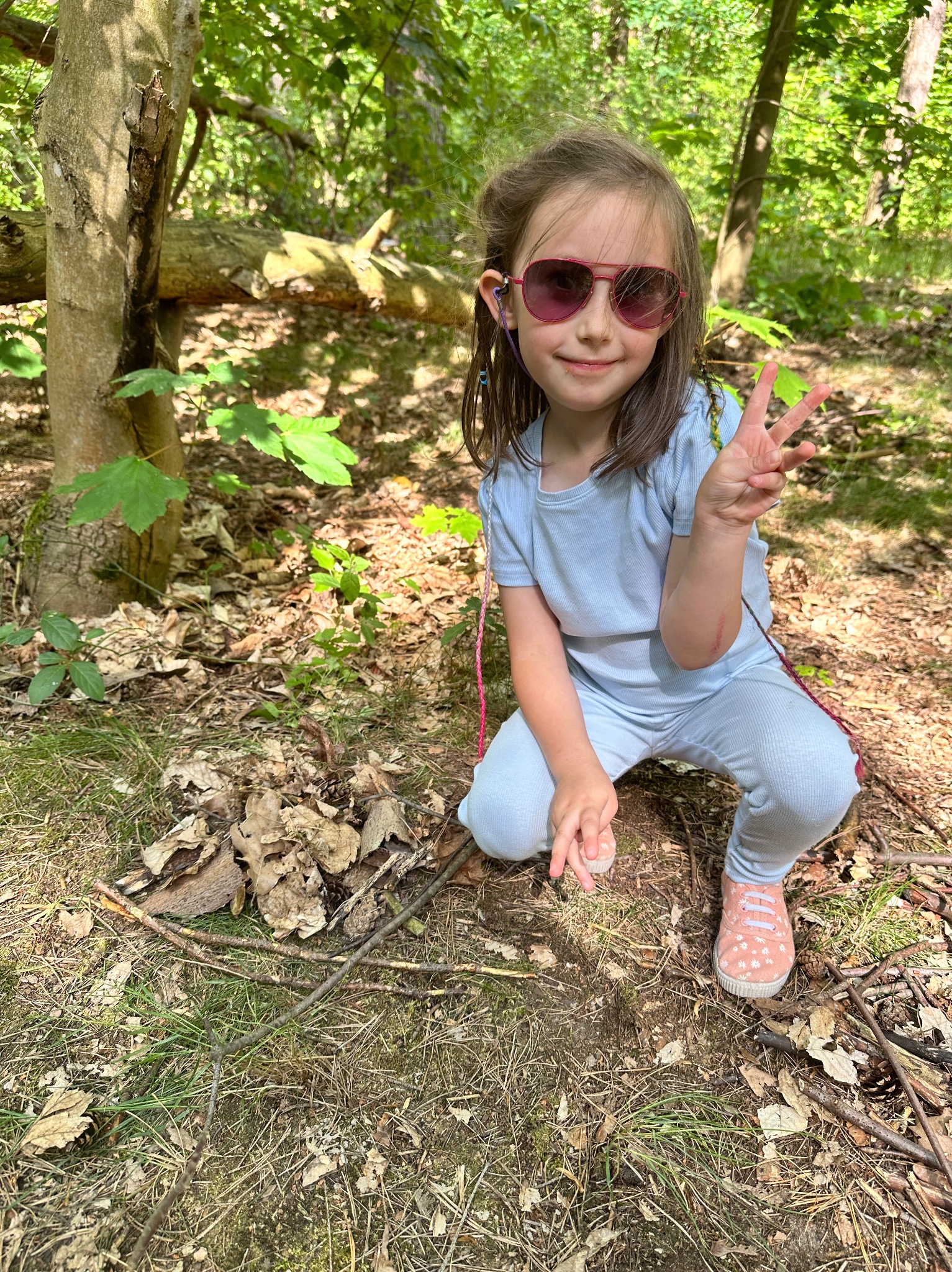 Uśmiechnięta dziewczynka w okularach i błękitnym dresie, kuca w lesie, pokazując palcami znak zwycięstwa. W tle drzewa i leśne poszycie.