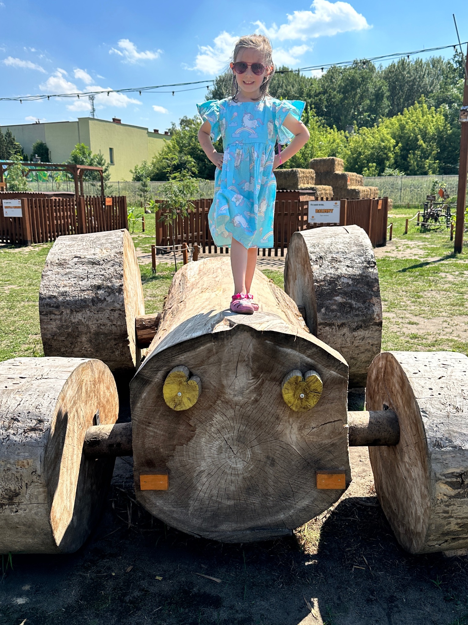 Dziewczynka w niebieskiej sukience w jednorożce stoi na drewnianej konstrukcji imitującej traktor z pni drzew, na tle zieleni i ogrodzenia.