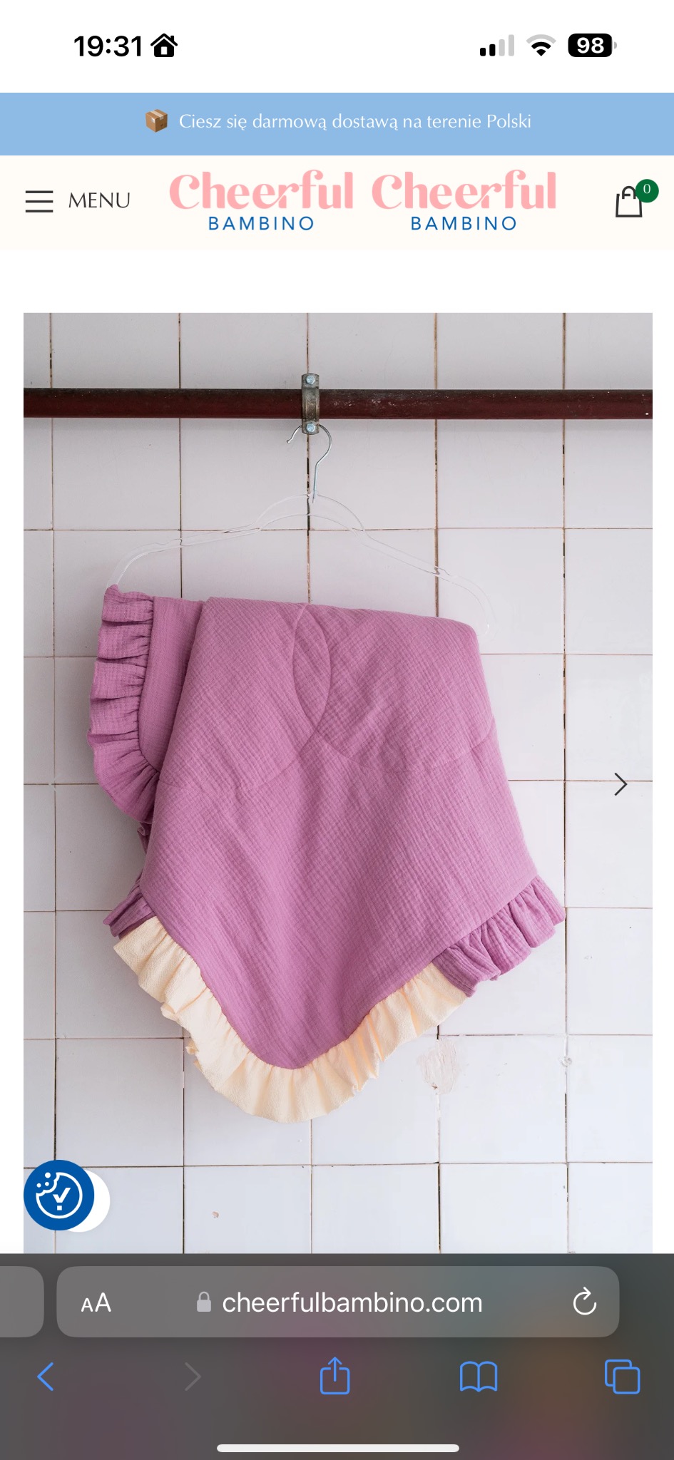 Różowy kocyk dziecięcy z kremowymi falbankami, zawieszony na wieszaku na tle białych płytek.