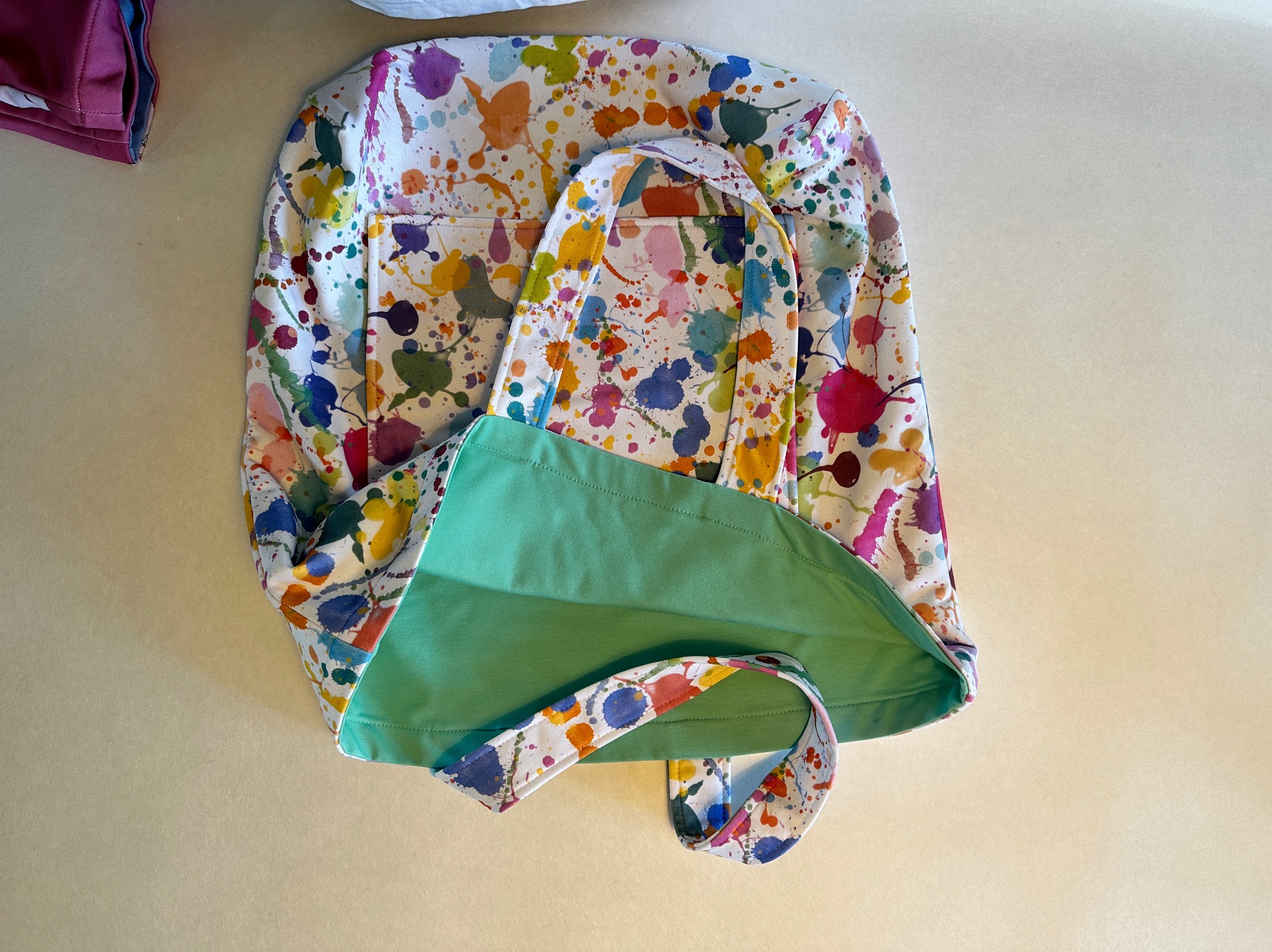 Otwarta torba na zakupy z kolorowym wzorem plam farby i zielonym wnętrzem, leżąca na jasnym tle.