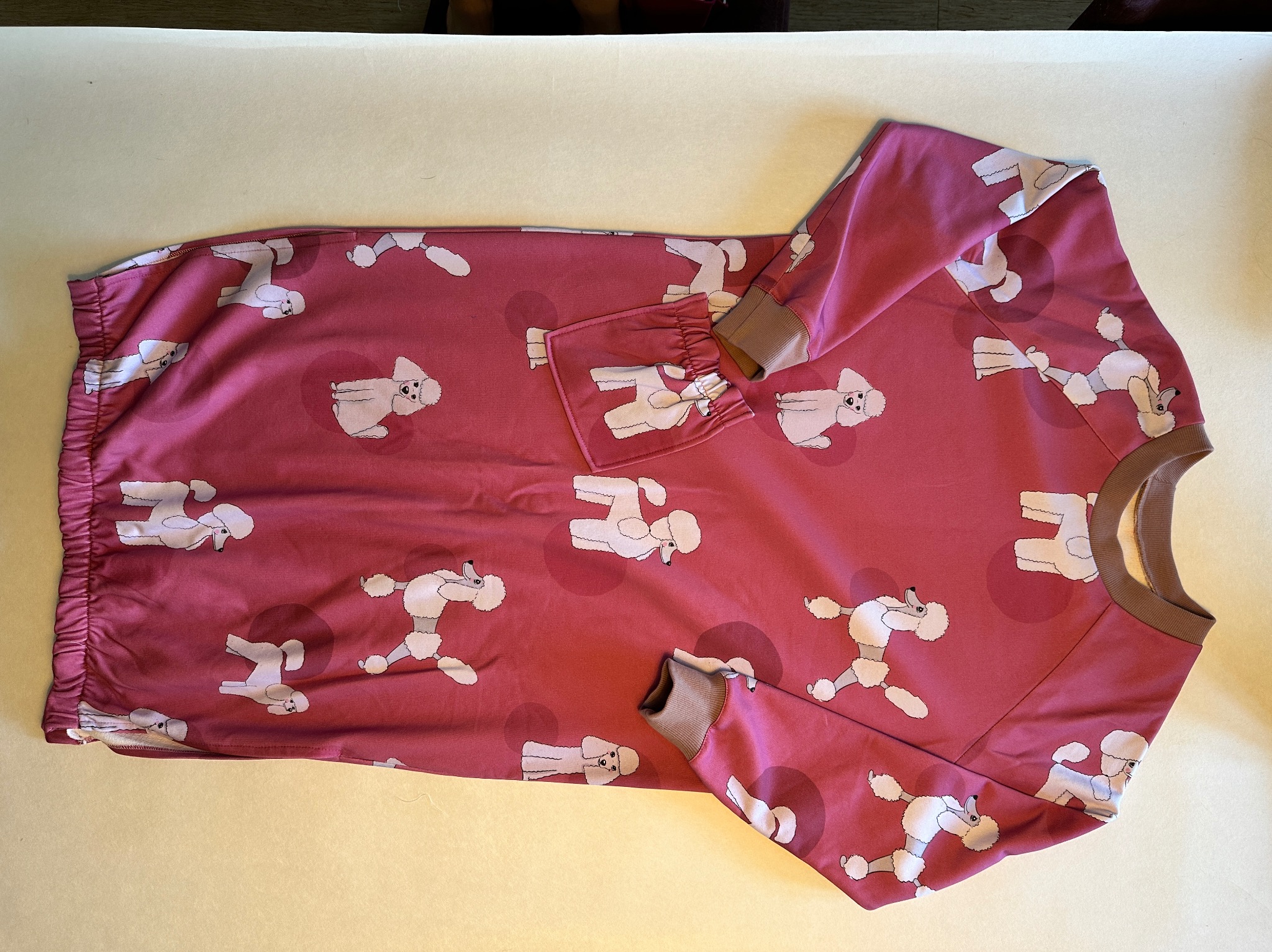 Dziecięca bluza w kolorze malinowym z motywem białych pudli, wykończona brązowymi ściągaczami przy szyi i rękawach, leżąca na jasnym tle.