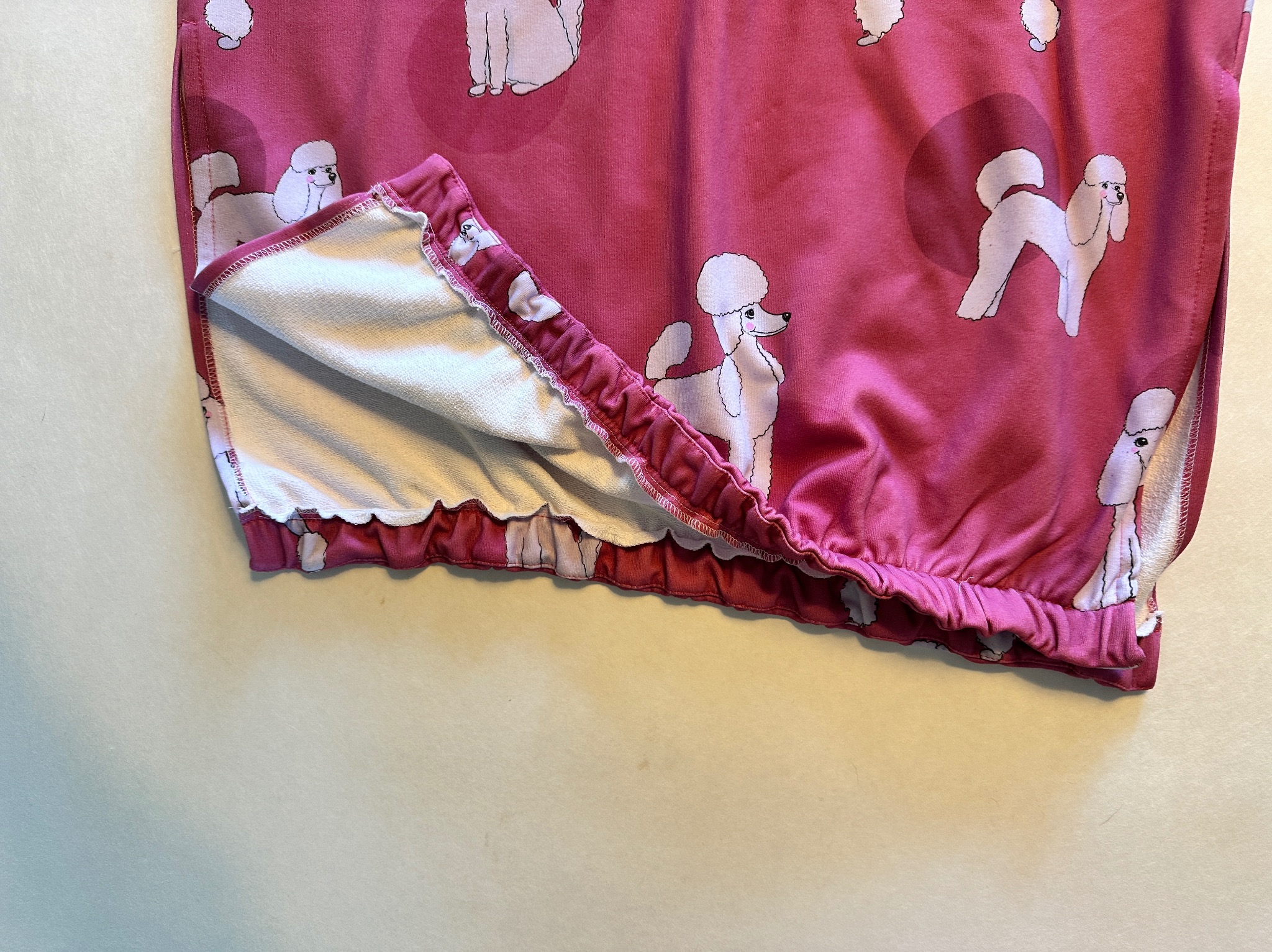 Fragment różowej dziecięcej bluzy z motywem białych pudli, wykończonej elastycznym ściągaczem, widoczne szwy i podszewka z białego materiału frotte.