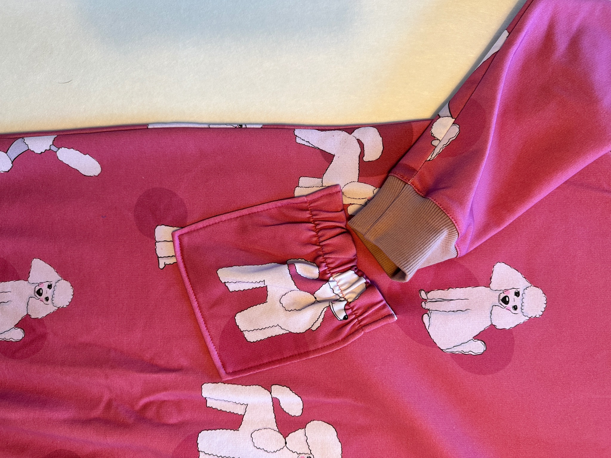 Fragment różowej bluzy z długim rękawem, pokrytej wzorem białych pudli, mankiet wykończony brązowym ściągaczem, widoczne marszczenia przy zakończeniu rękawa.